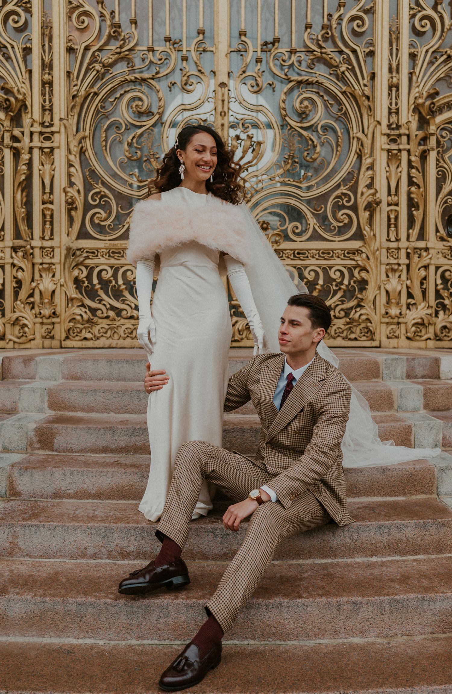 Stylish couple in vintage-inspired wedding attire pose on stone steps with ornate gold gates.
