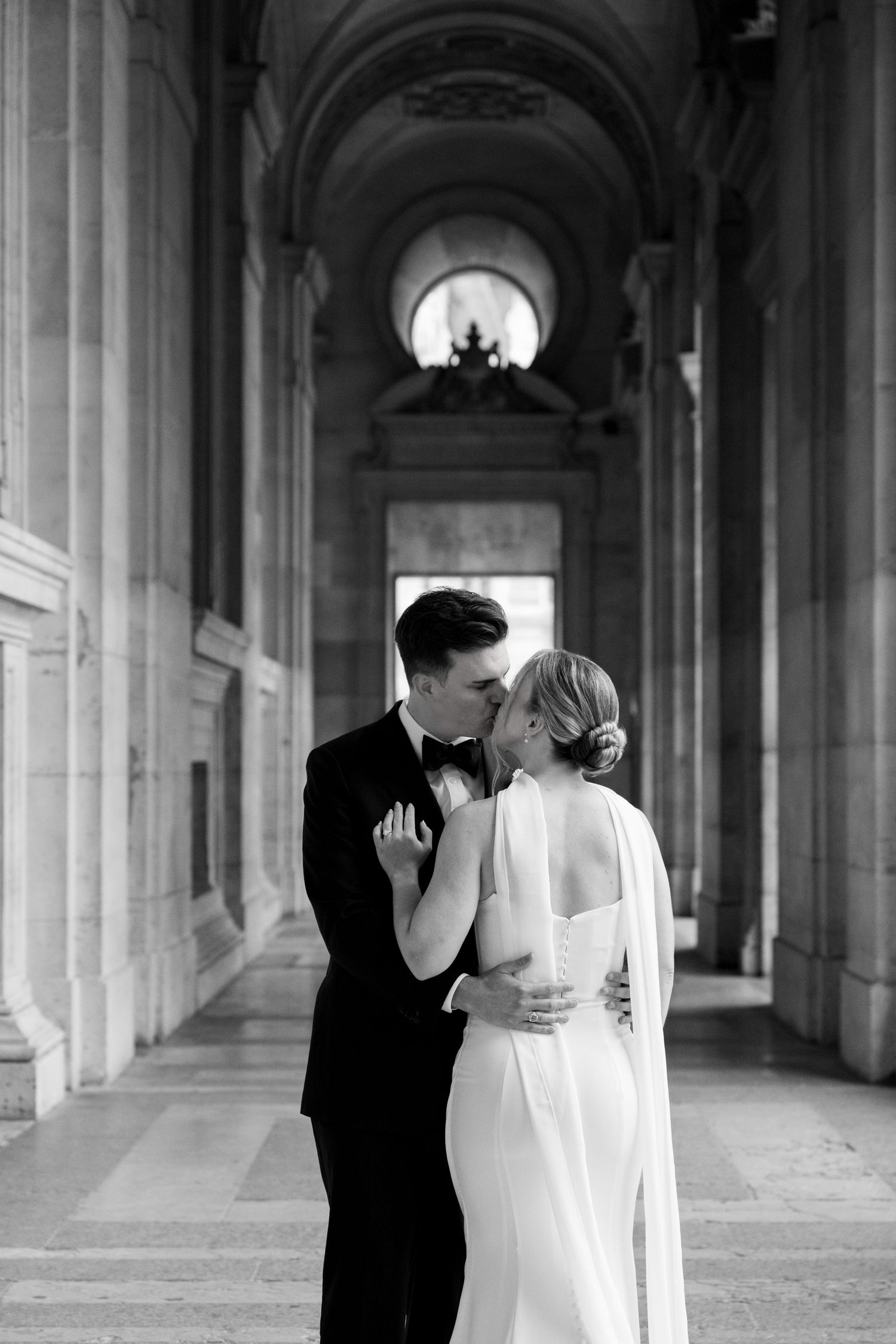 A couple shares an intimate moment in an elegant arched hallway with classical architecture.