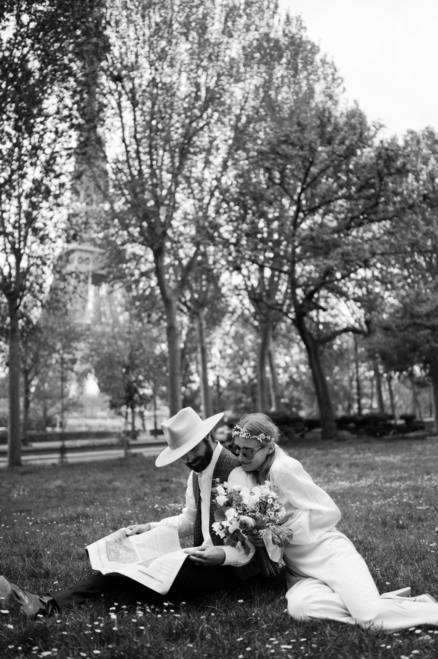 Black and white photo of couple reading in park with bouquet and trees.