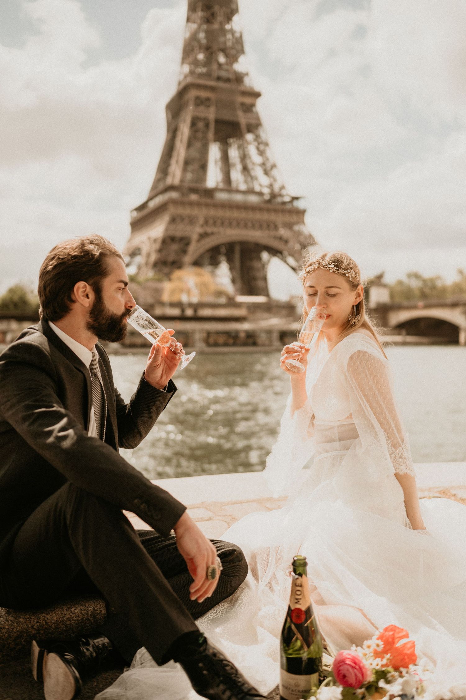 Newlyweds sharing champagne and laughter by the Seine with Eiffel Tower in background.