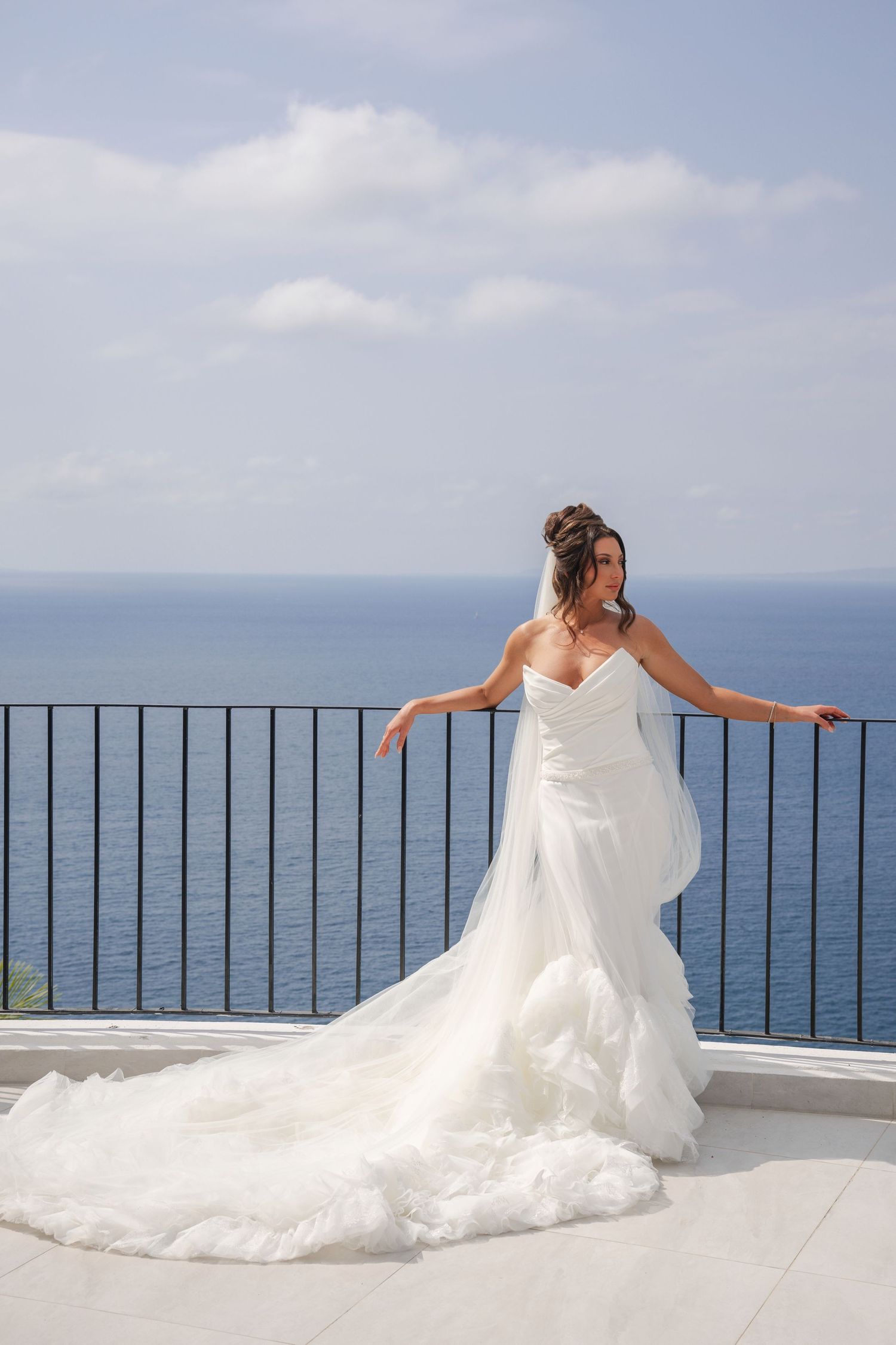 Beautiful bride in a ruffled white wedding dress leans against a balcony railing with sea backdrop.