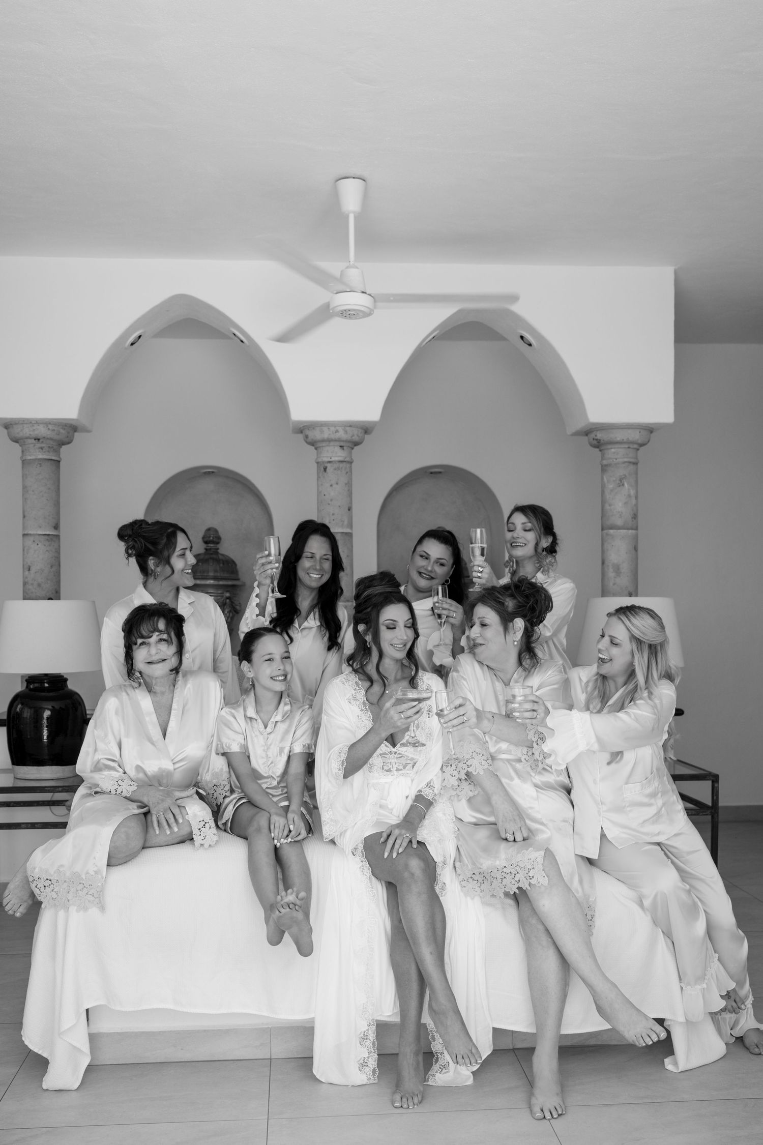 Bridesmaids in white satin robes toasting with champagne glasses in a black and white wedding preparation photo.