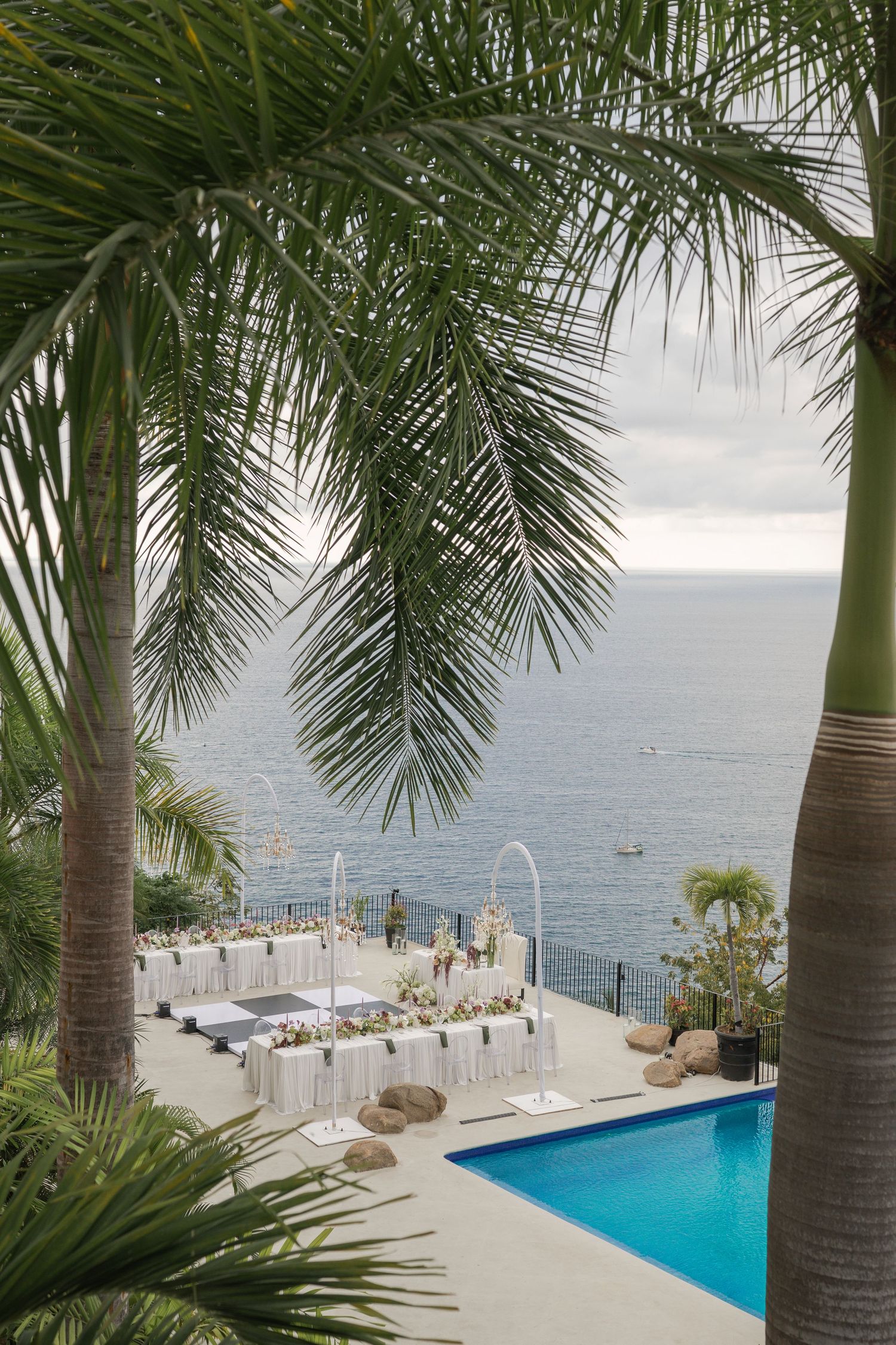 Outdoor oceanfront wedding reception setup with white tables near a pool, framed by tropical palm trees.