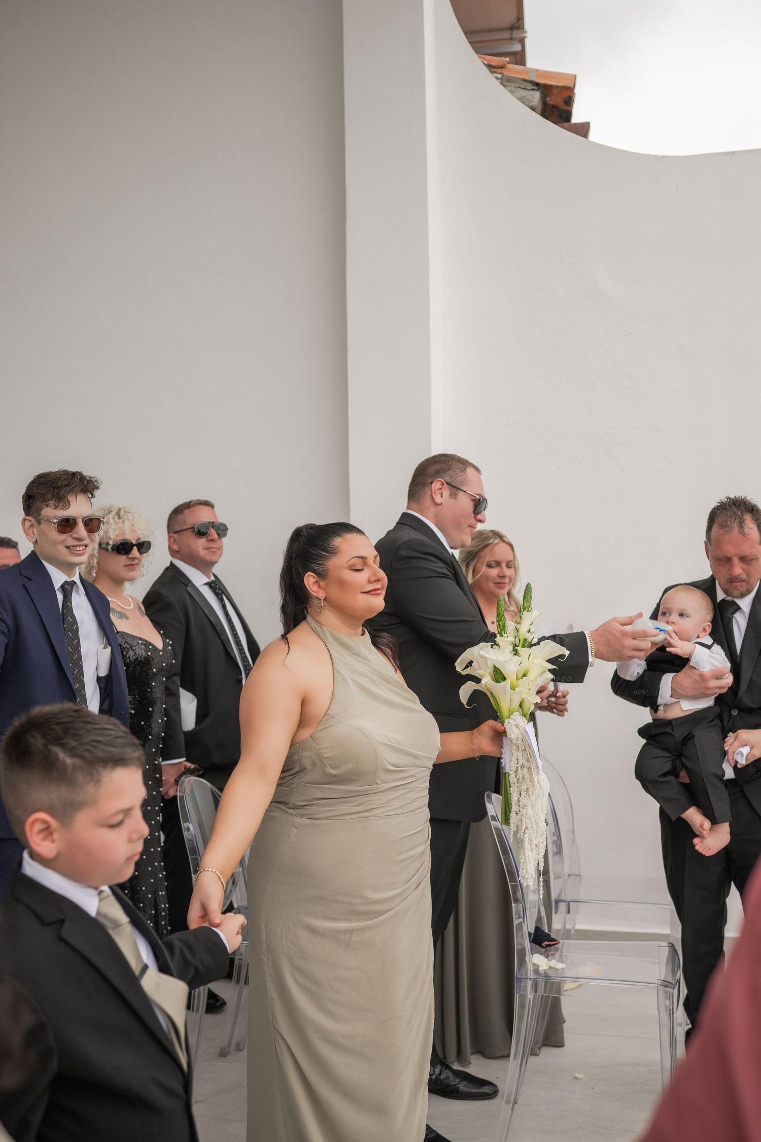 Wedding guests gathered indoors as bridesmaid smiles holding white lily bouquet.