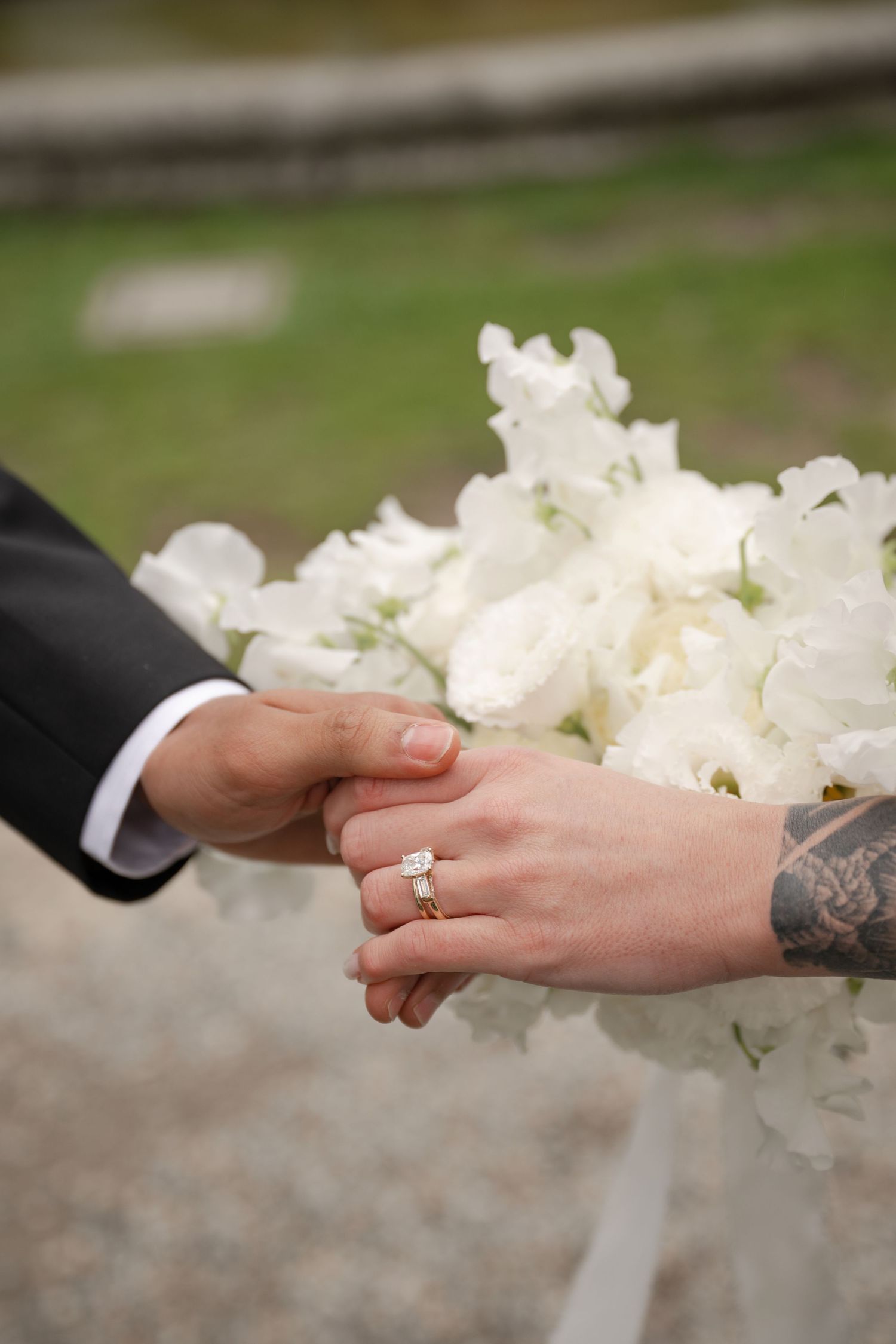 Bride and groom holding hands with diamond engagement ring and tattoo visible, white floral bouquet in background.