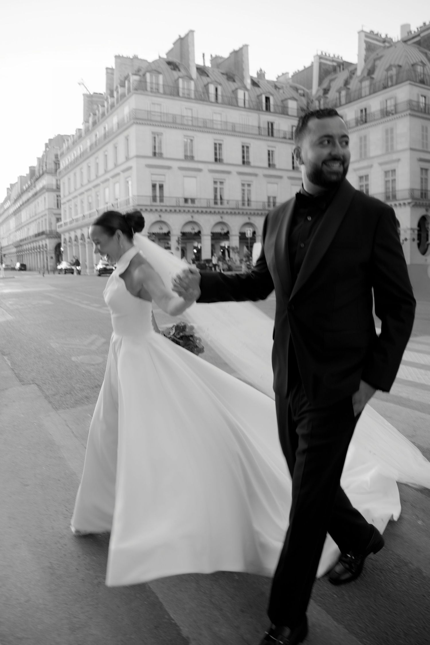 Black and white photo of a smiling groom in a suit leading his bride in a flowing white dress through Paris streets.