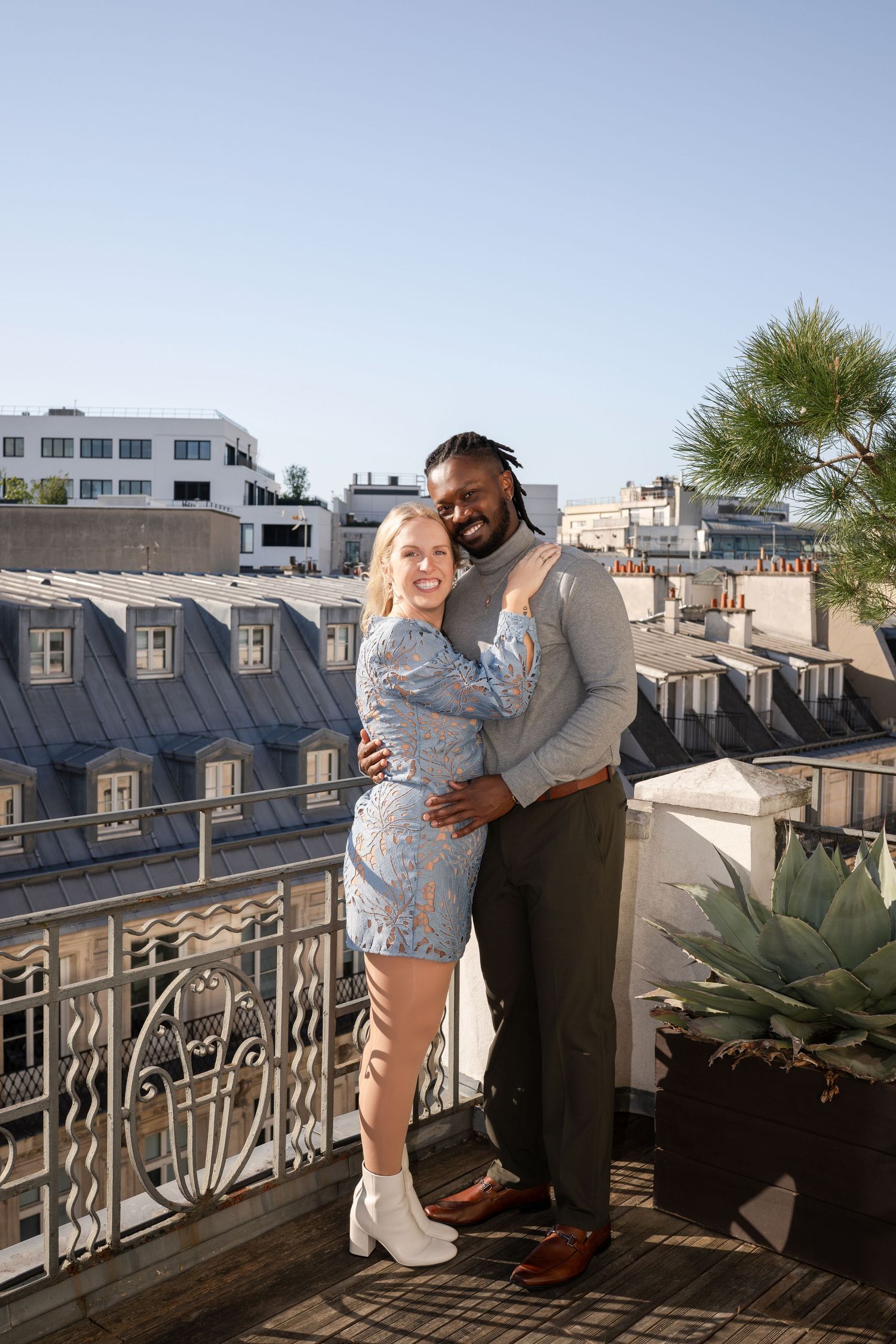 A happy interracial couple hugs and smiles on a sunny Parisian rooftop terrace with Haussmann buildings behind them.