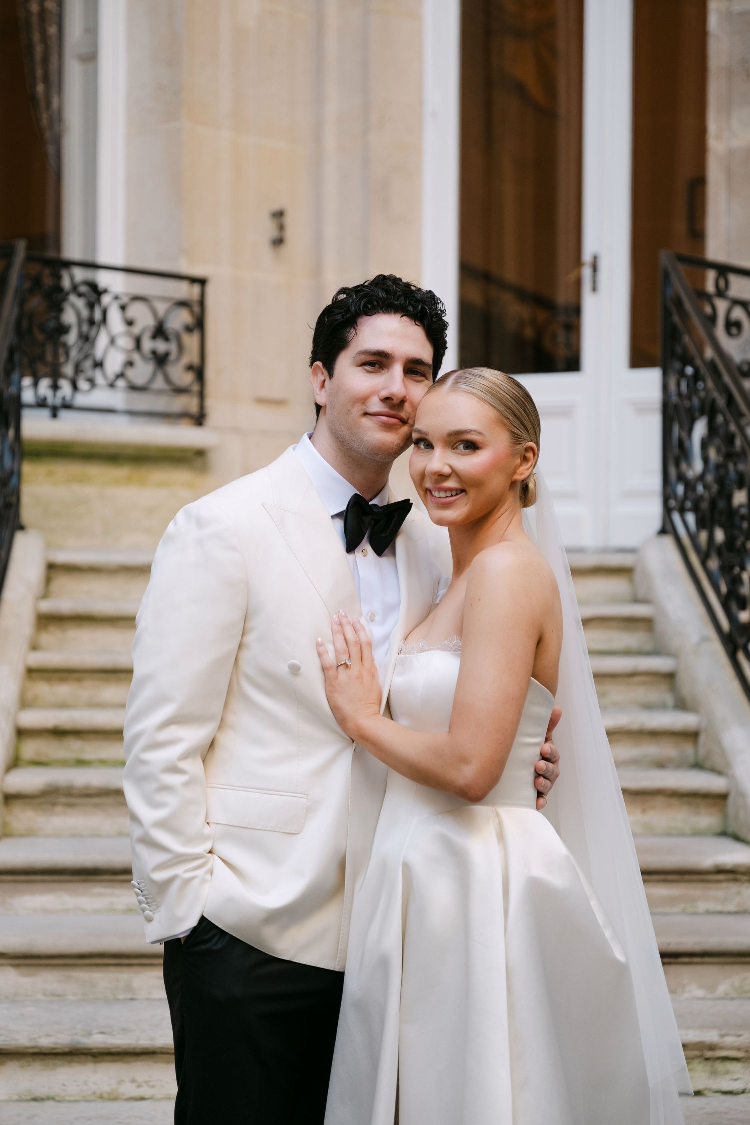 Smiling newlyweds embrace on steps of French chateau, groom in white tuxedo jacket with black bow tie.