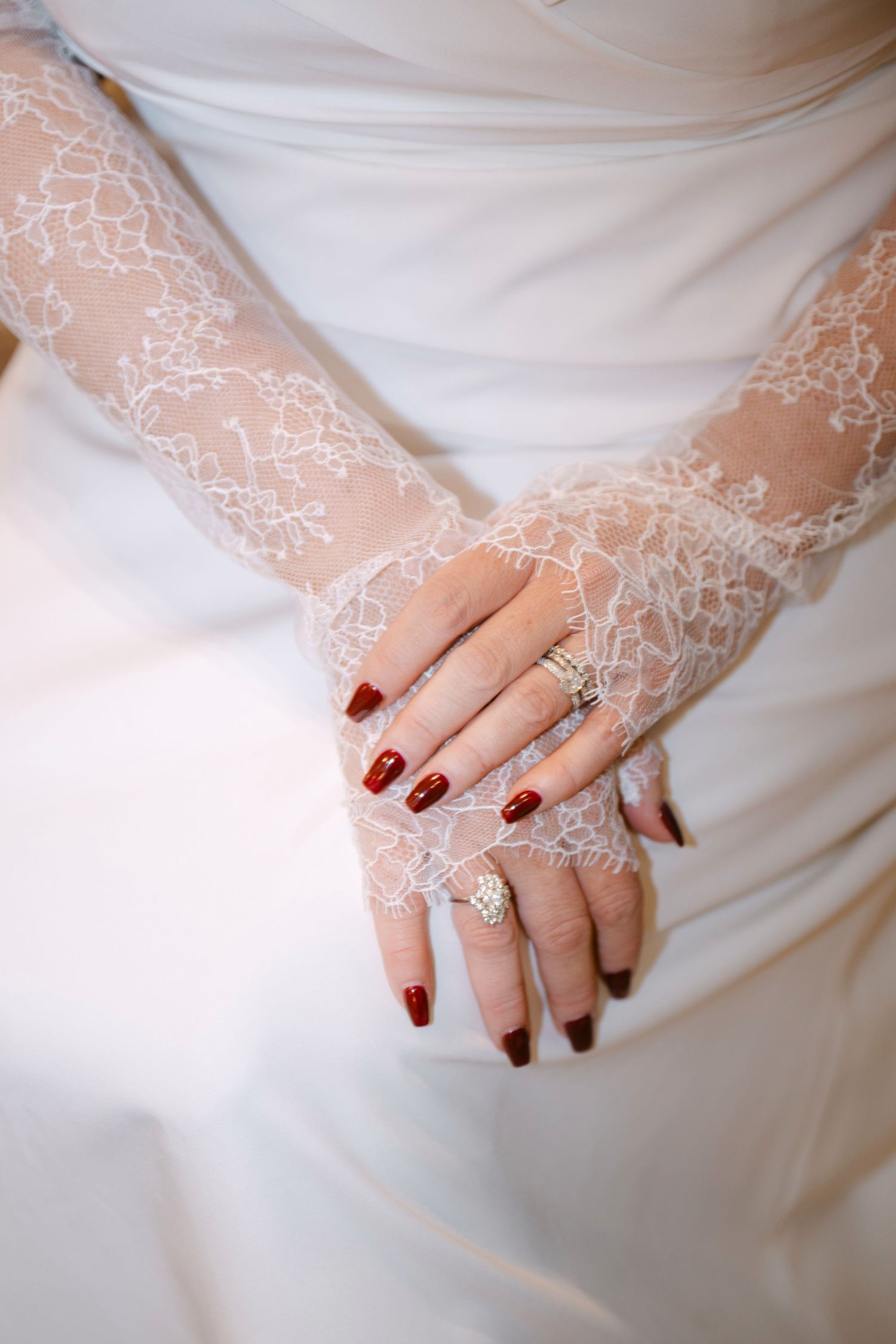 Bride wearing lace fingerless gloves with dark red nails and diamond rings on white wedding dress.