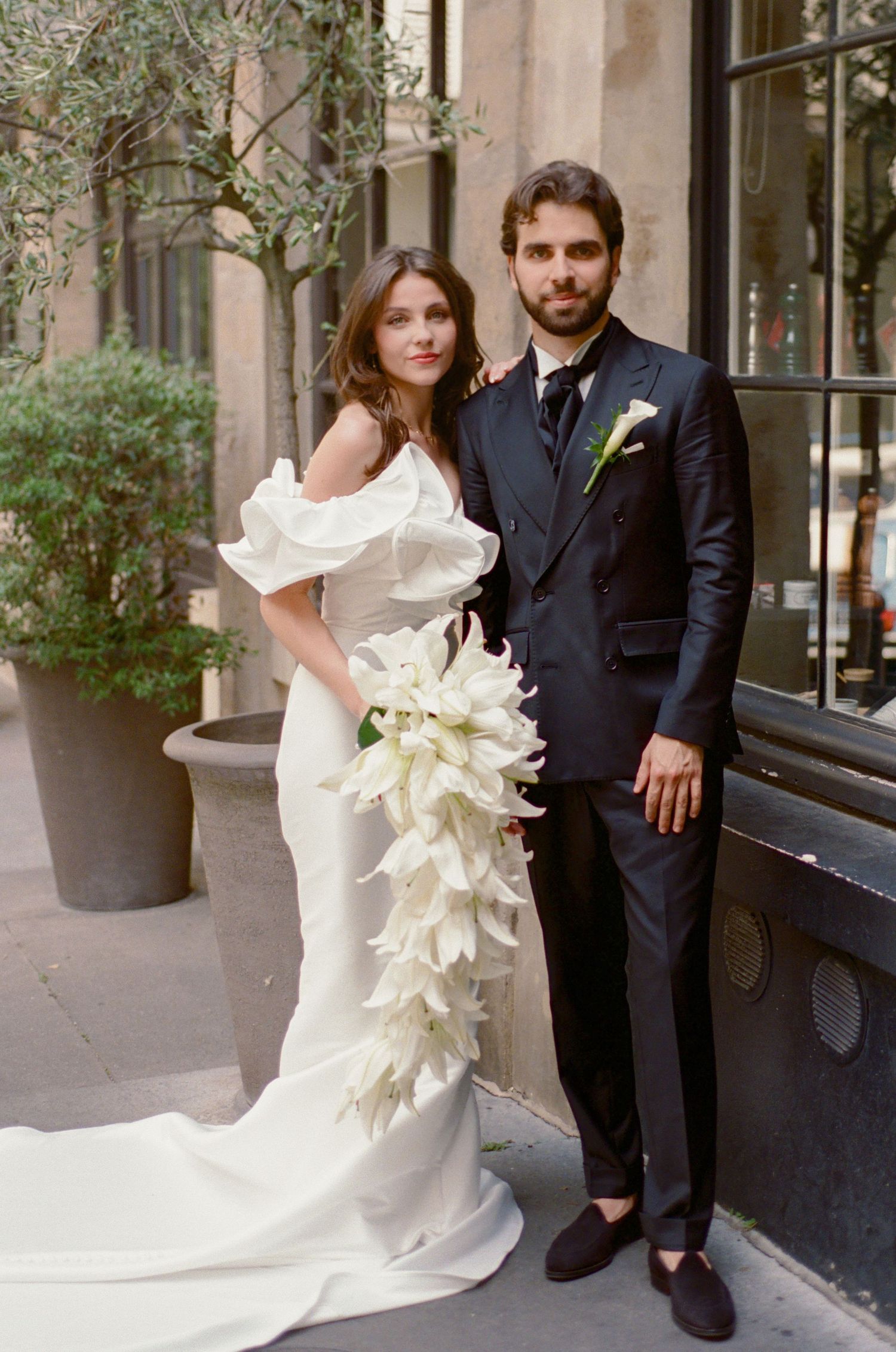 Bride in off-shoulder white gown holding cascading white lily bouquet stands with groom in dark suit on city sidewalk.