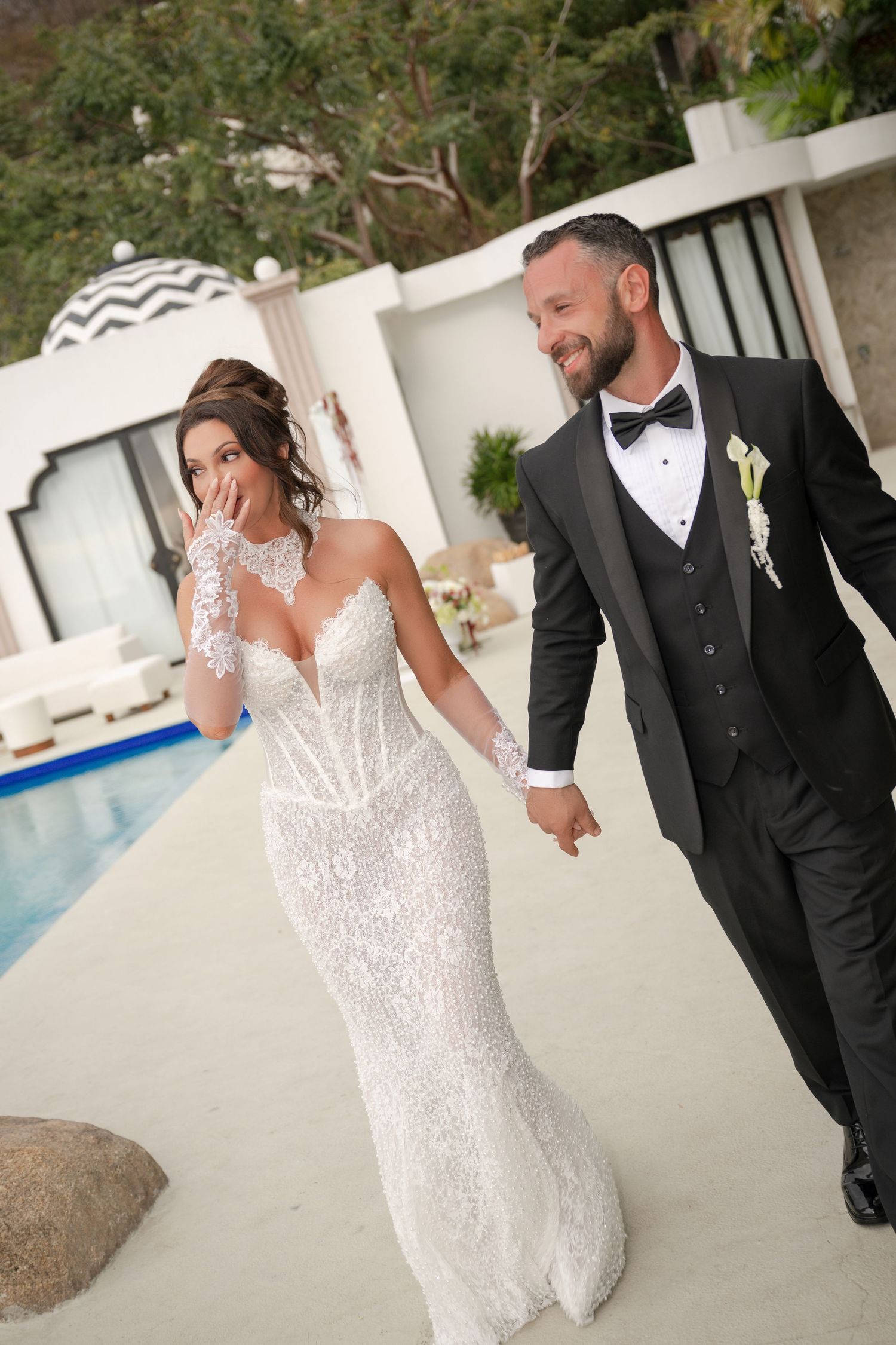 Smiling bride and groom in elegant wedding attire walking hand in hand by outdoor pool.
