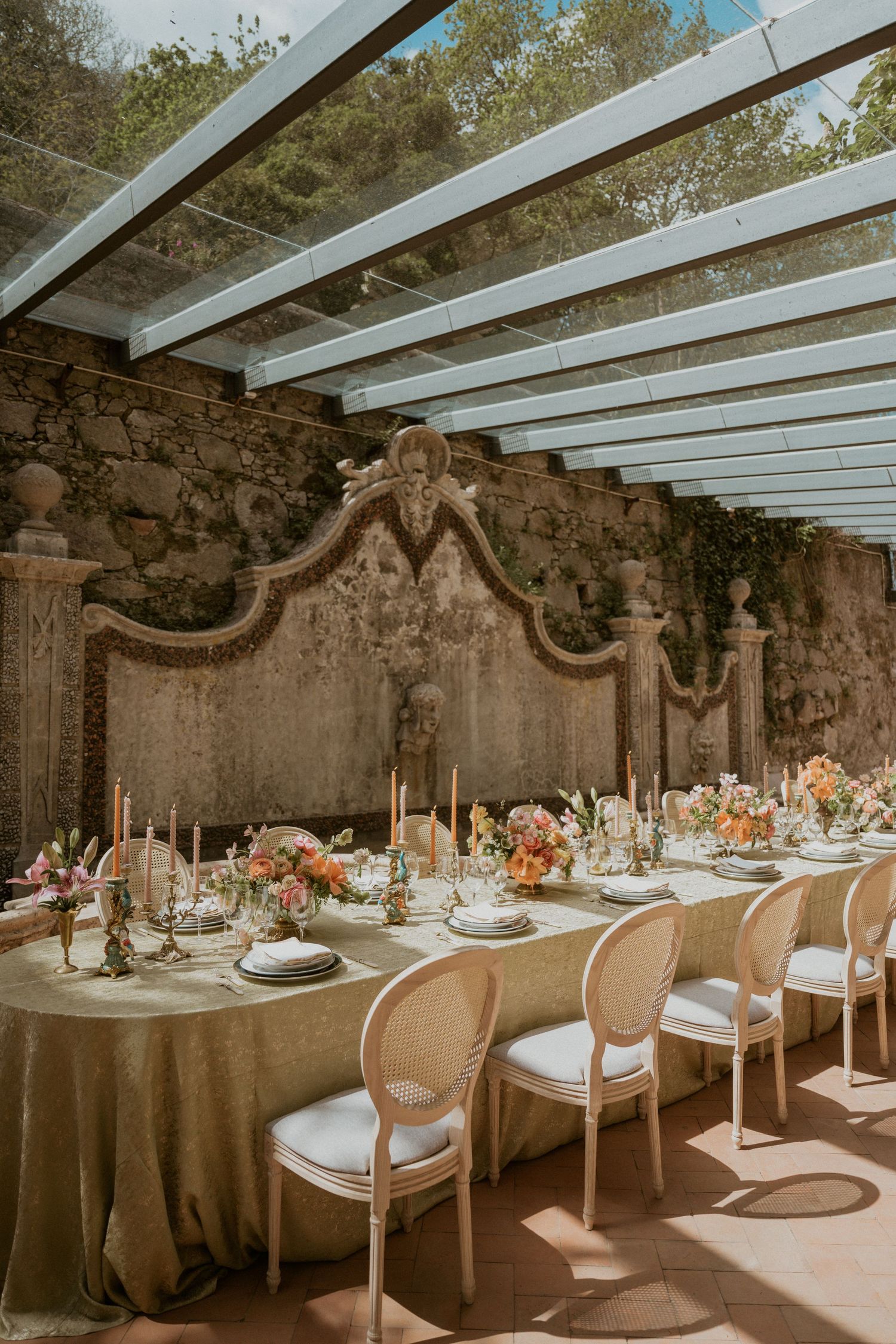 Elegant outdoor wedding reception with long table, gold linens, floral centerpieces, and Louis XVI chairs against a stone wall.