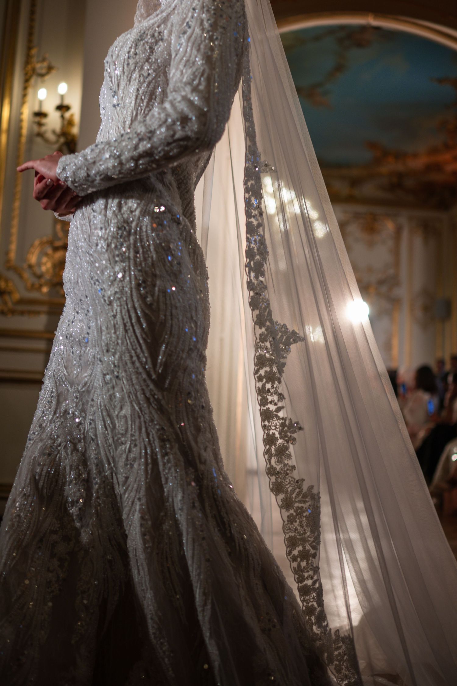 Bride in an ornate beaded mermaid wedding gown with long sleeves and embroidered veil in an elegant ballroom.
