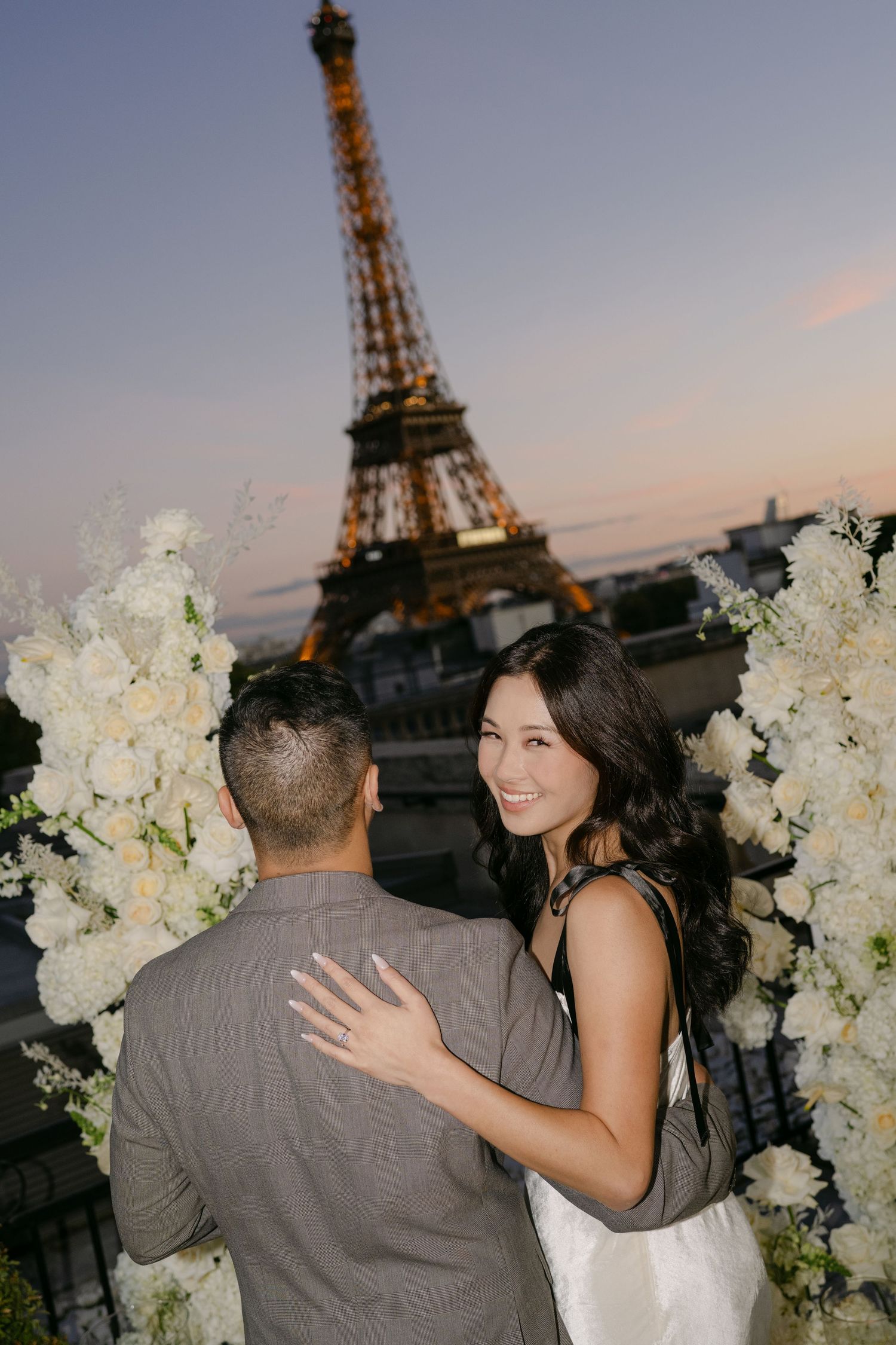 Smiling woman showing her ring while embracing a man on a floral-decorated Paris rooftop with the Eiffel Tower glowing at sunset.