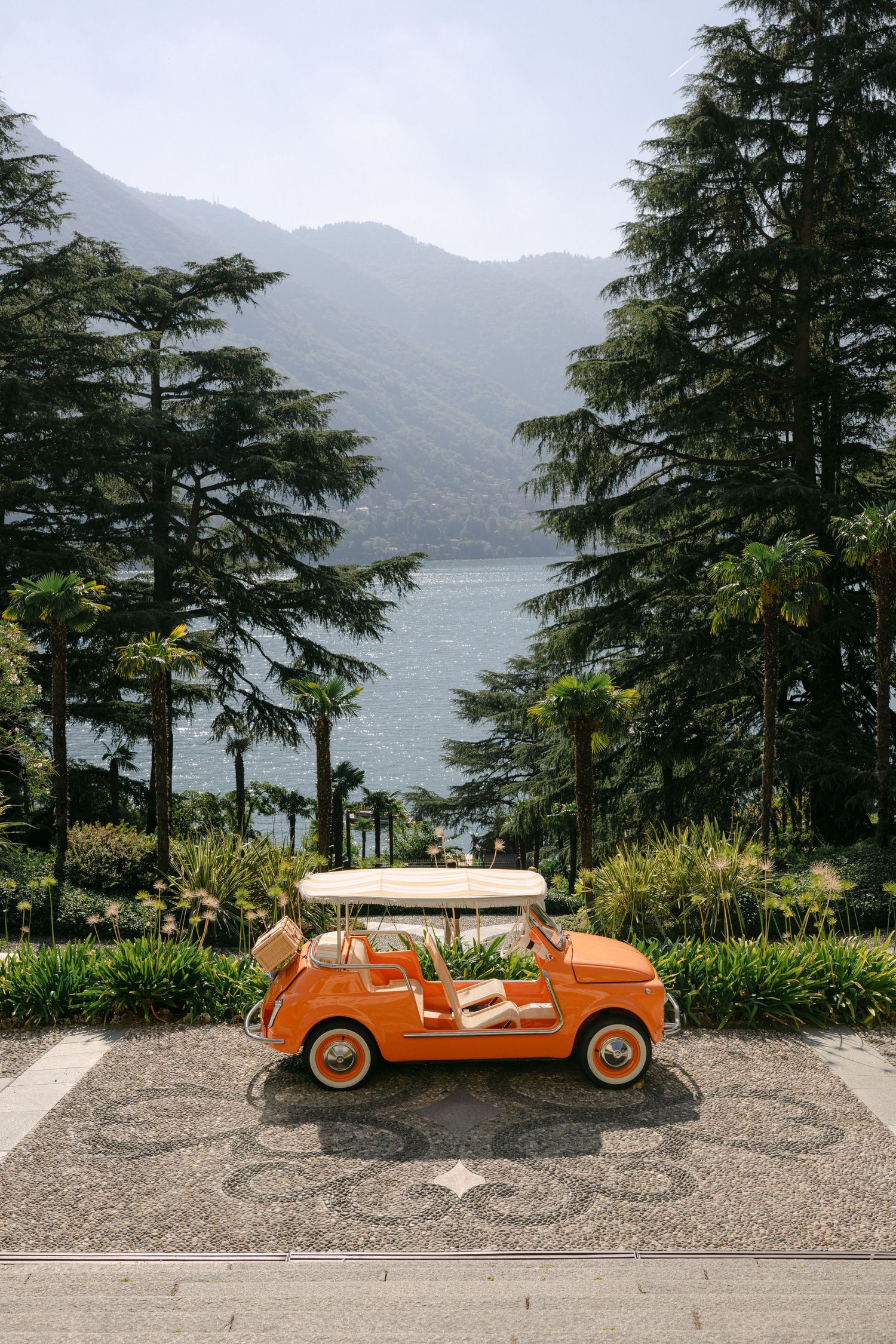 Vintage orange Fiat 500 convertible parked on mosaic driveway with Lake Como and mountain backdrop in Italy.