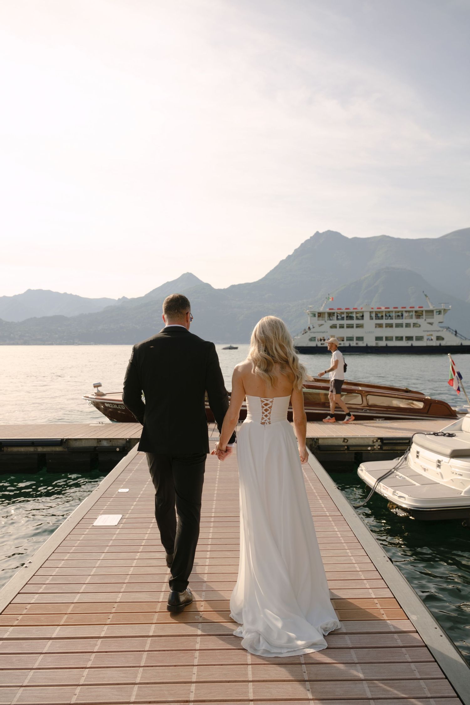 Bride and groom walking hand in hand on a wooden dock with mountains and a ferry in the background.