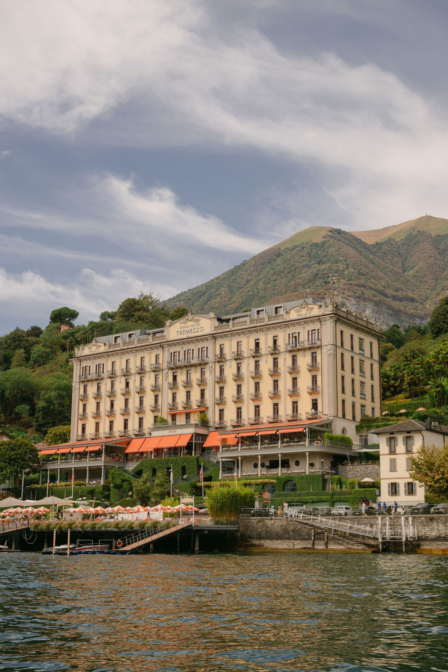 Grand hotel on Lake Como waterfront with red awnings, lush mountain backdrop, and cloudy sky in Italy.