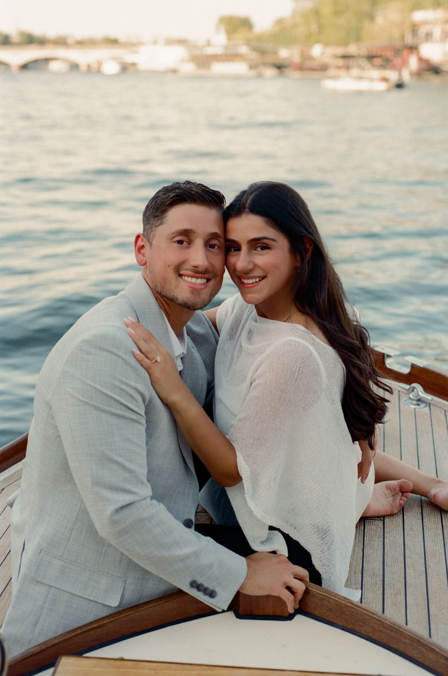 Happy couple embracing on a wooden boat with a scenic waterway and bridge in the background.
