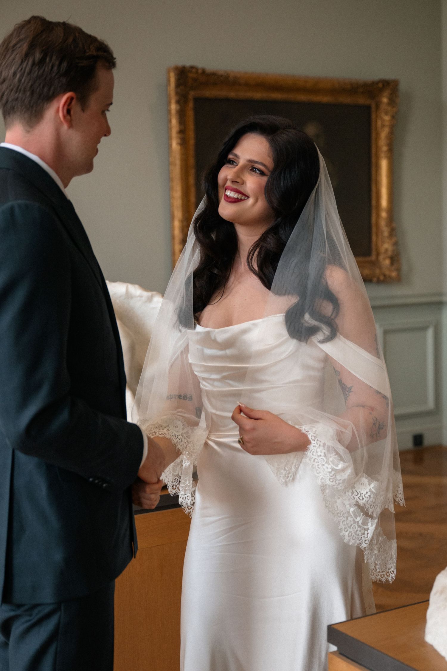 Smiling bride in satin wedding dress and lace veil holds grooms hands before a golden framed painting.
