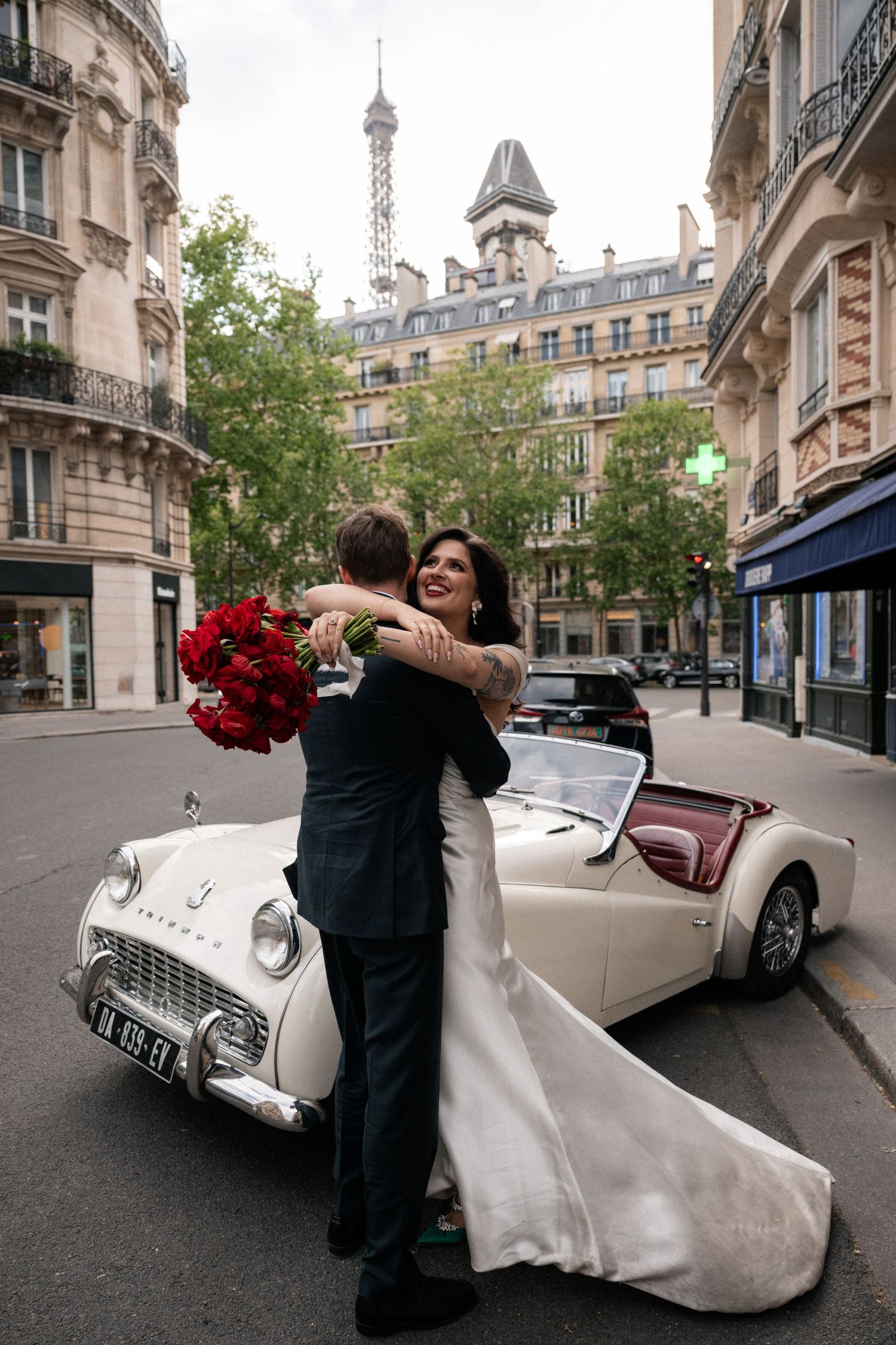 Joyful bride hugging groom beside classic white Triumph convertible with Eiffel Tower visible in background.