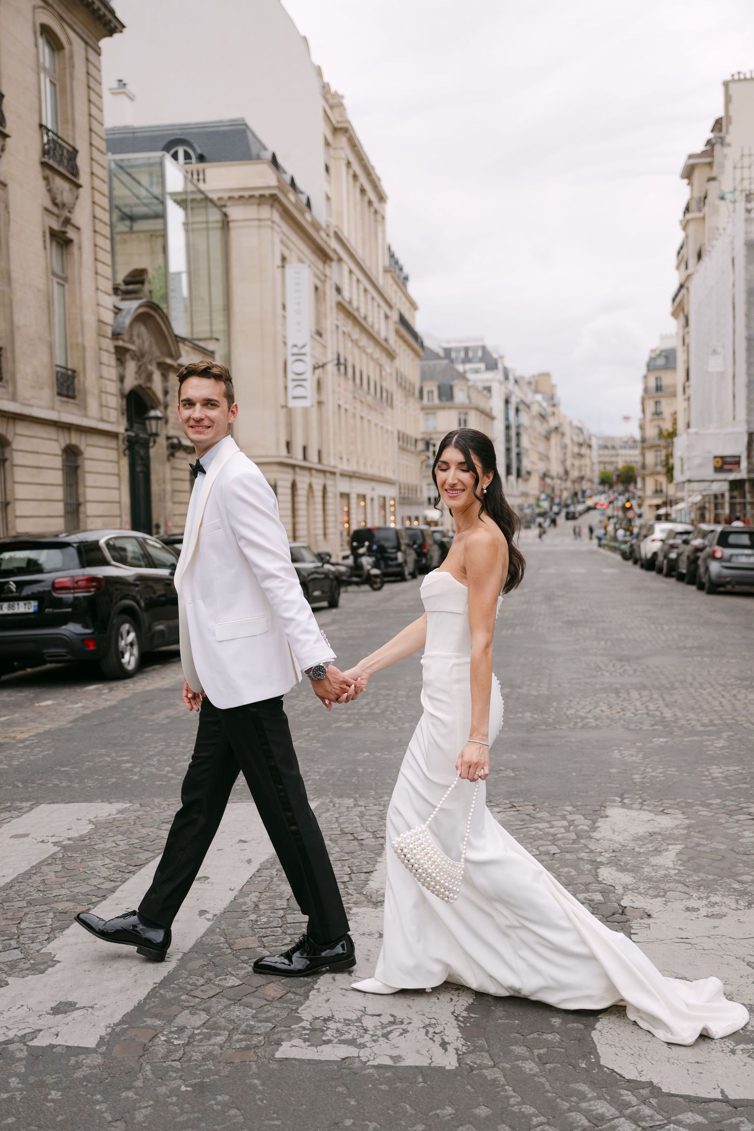 Bride and groom hold hands while crossing a classic Parisian boulevard, smiling at each other.