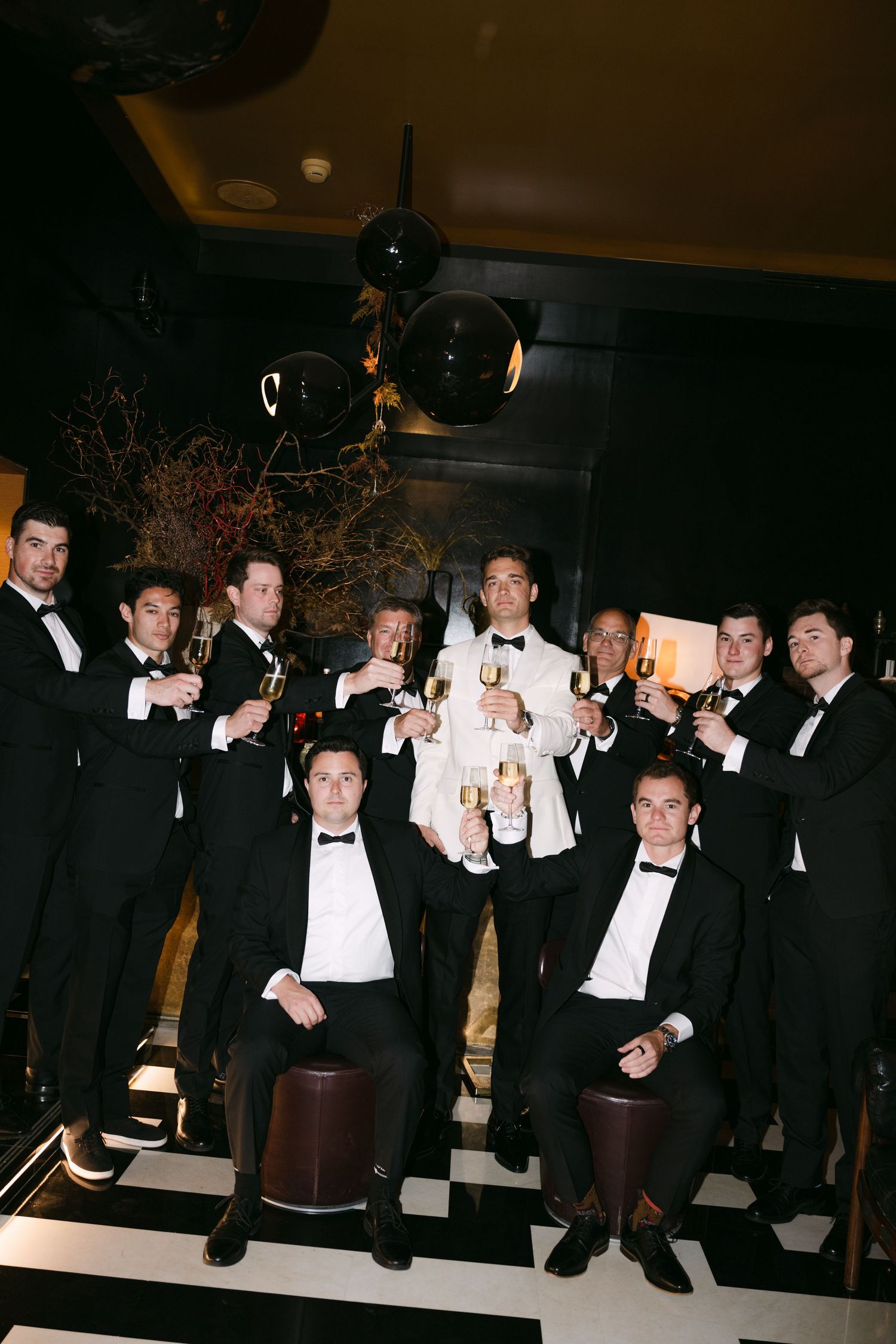 Groomsmen in black tuxedos toast champagne with groom in white jacket at sophisticated wedding celebration.