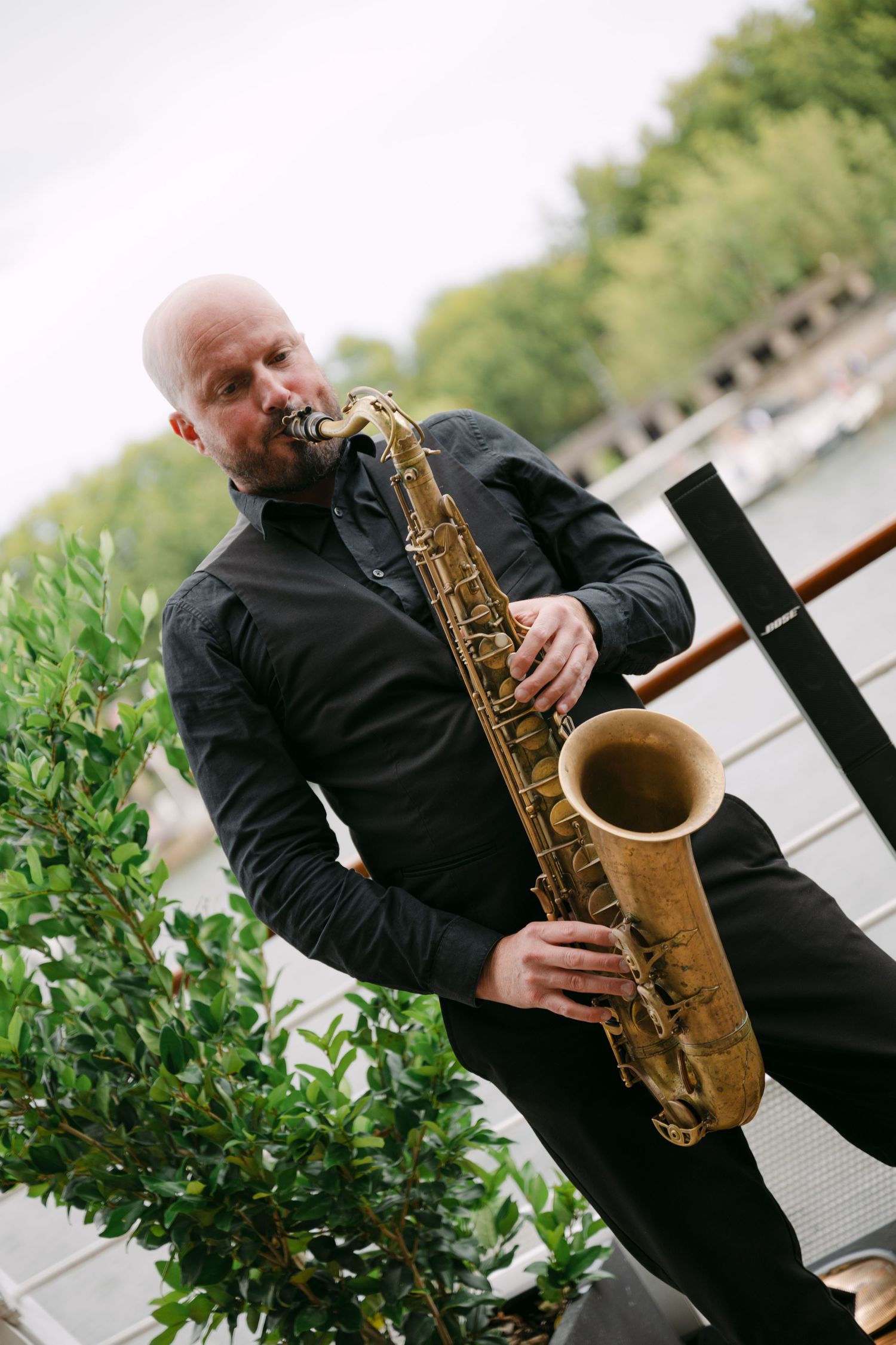 Bald saxophonist in black shirt performs outdoors near a river at a wedding reception in Paris.
