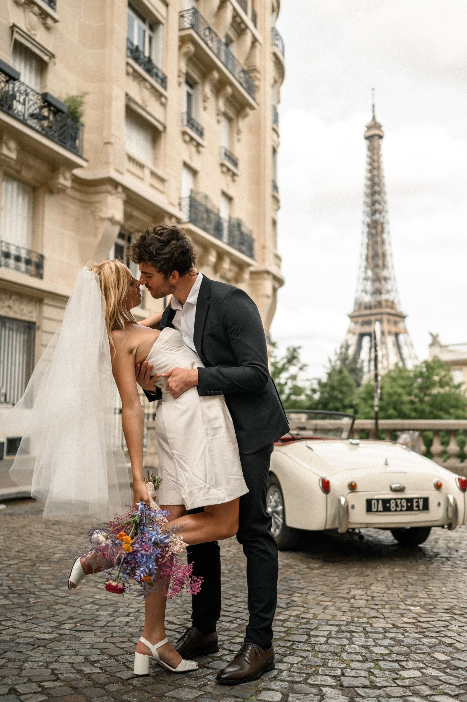 Bride and groom sharing romantic kiss in Paris with Eiffel Tower and vintage white Triumph convertible behind them.