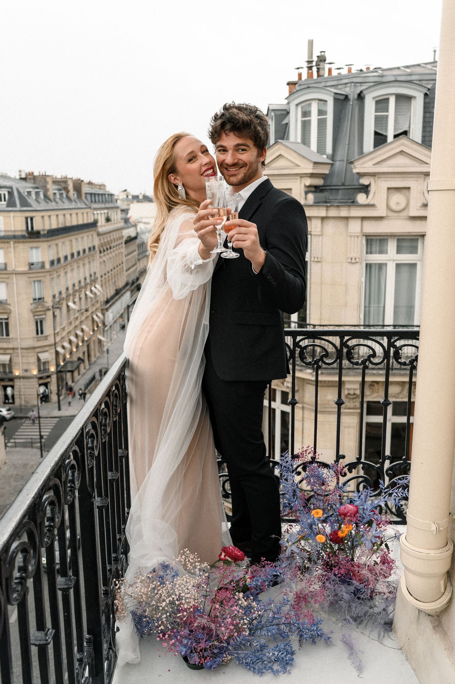 Newlyweds toasting champagne on a Paris balcony surrounded by colorful floral arrangements.