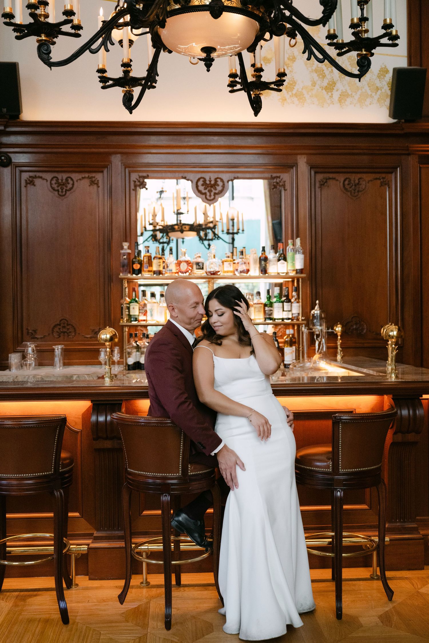 Bride and groom share intimate embrace at elegant wood-paneled bar under ornate chandelier.