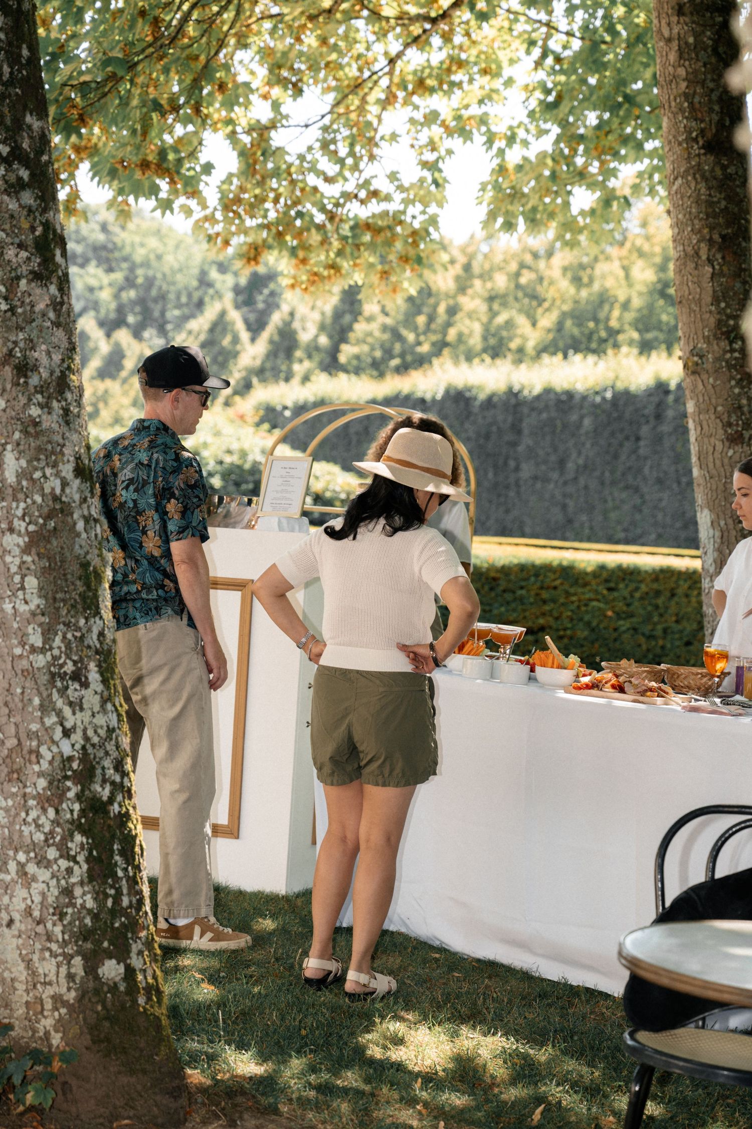 Guests browsing an outdoor catering table under trees at a sunlit garden wedding reception.