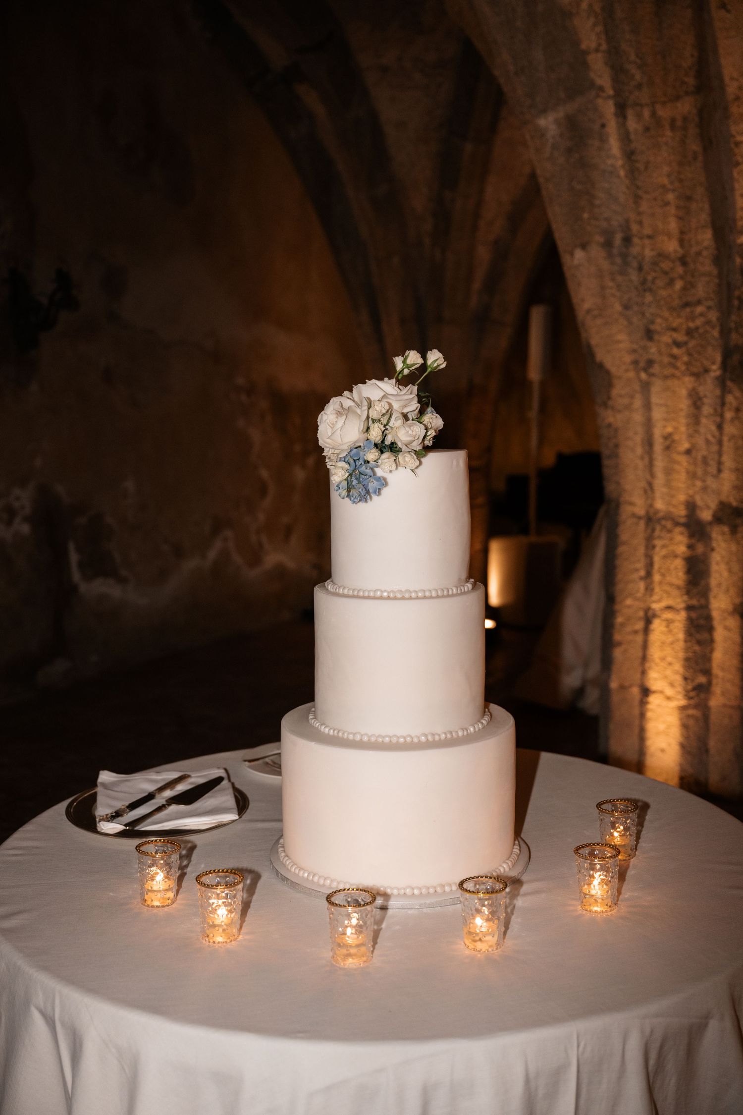 Three-tier white wedding cake adorned with white and blue flowers, surrounded by glowing votive candles.