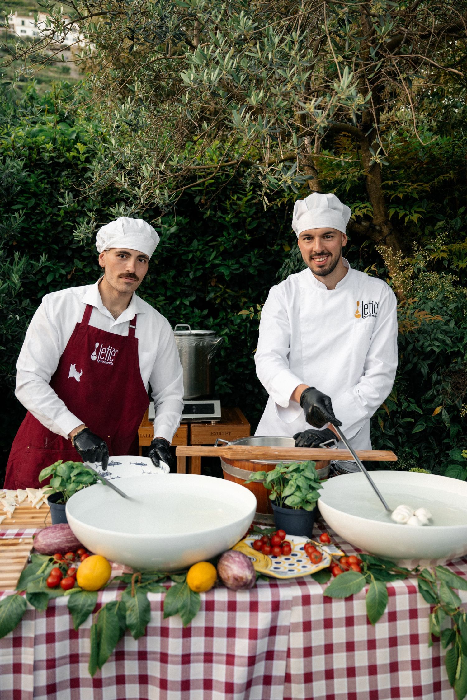 Two smiling chefs preparing fresh mozzarella and vegetables at an outdoor catering station.