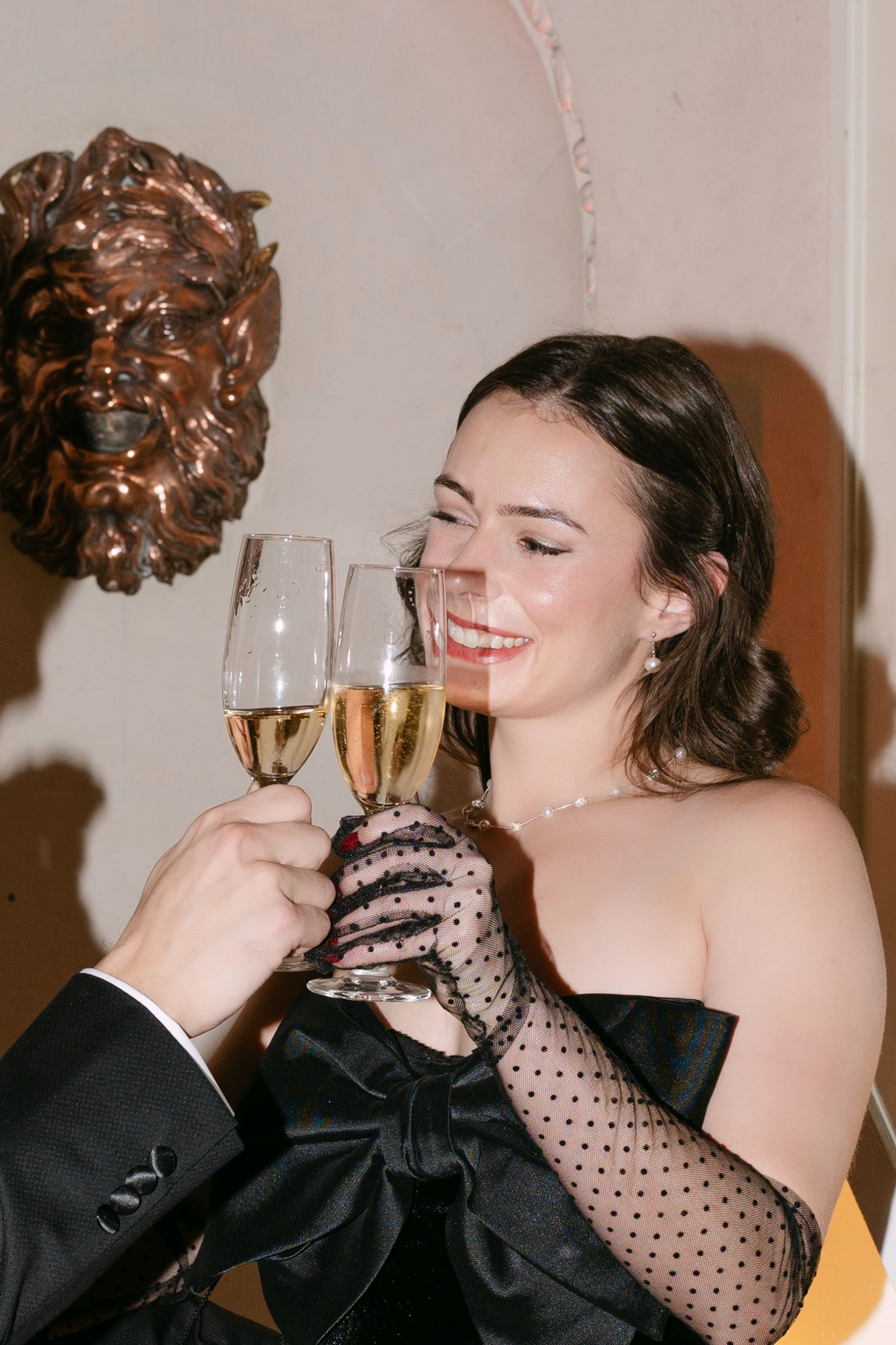 Smiling woman in black strapless bow gown with lace gloves toasting champagne flutes at elegant wedding celebration.