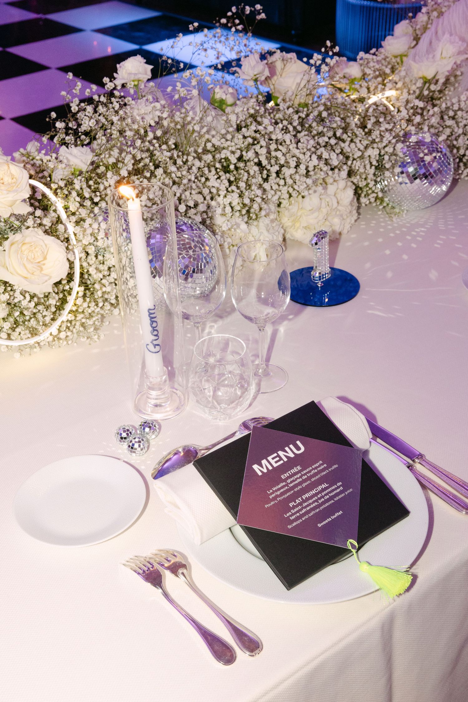 Wedding table setting with menu card, candle, crystal glasses, and white floral arrangements.