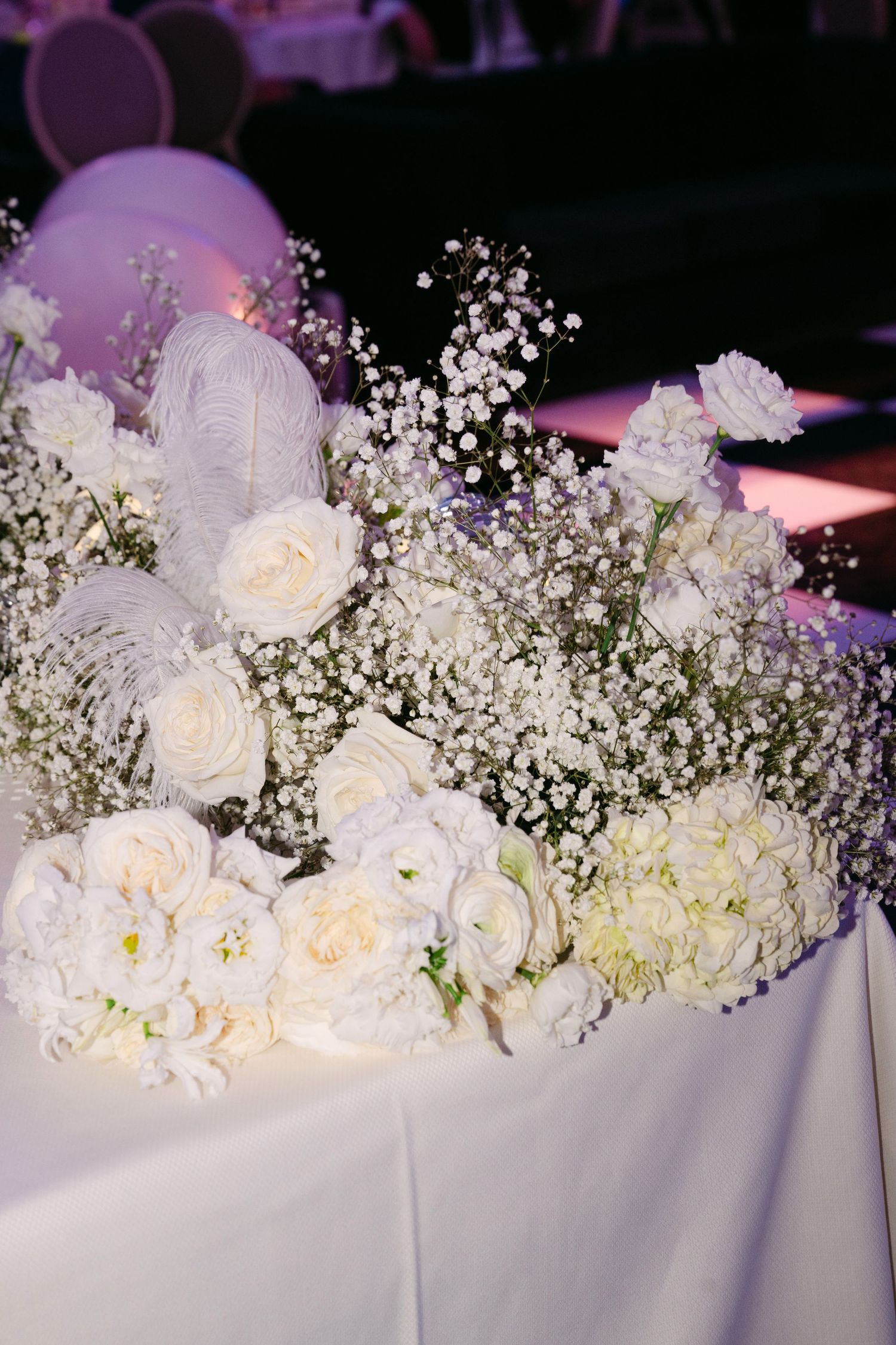 Lush white wedding centerpiece featuring roses, baby's breath, and feathers on reception table.