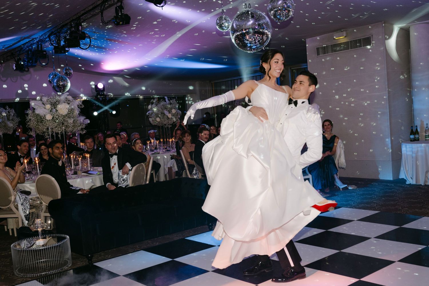 Groom lifts bride during their first dance on a black and white checkered dance floor.