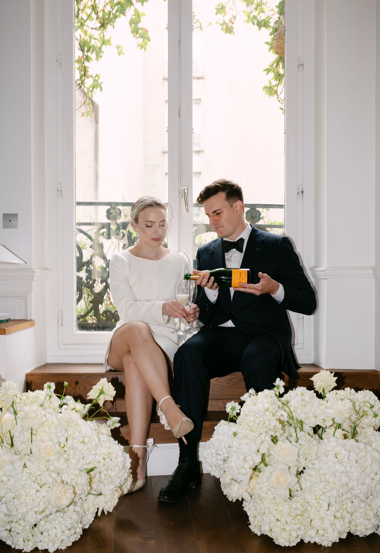 Elegant couple enjoys tea by window surrounded by white hydrangea flowers in romantic indoor setting.