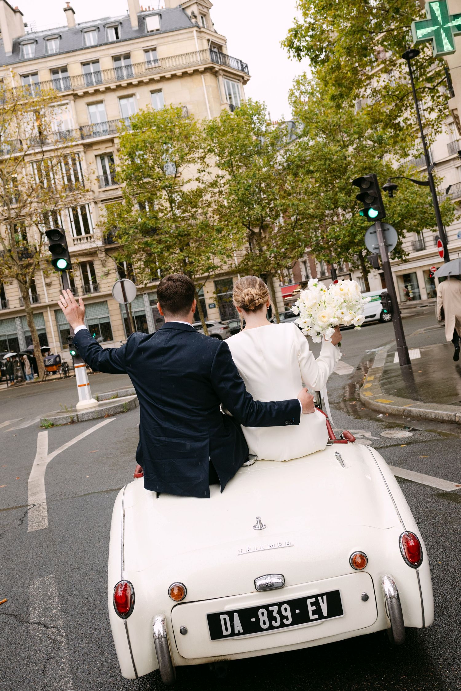 Newlyweds drive away in a vintage white convertible car through a charming Parisian street lined with classic buildings.