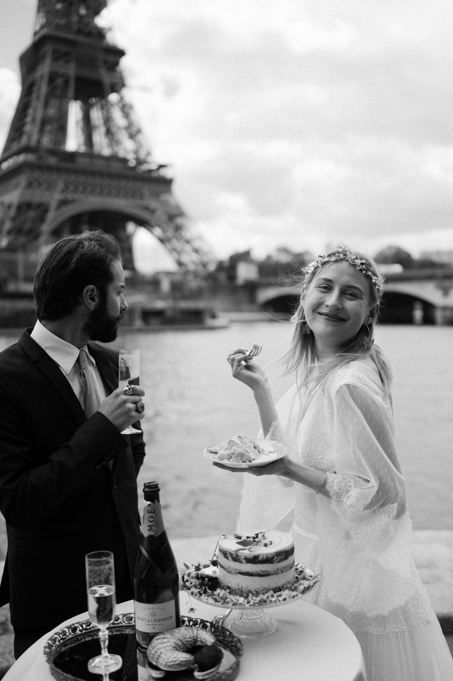 A black and white photo of newlyweds enjoying cake and champagne by the Eiffel Tower.