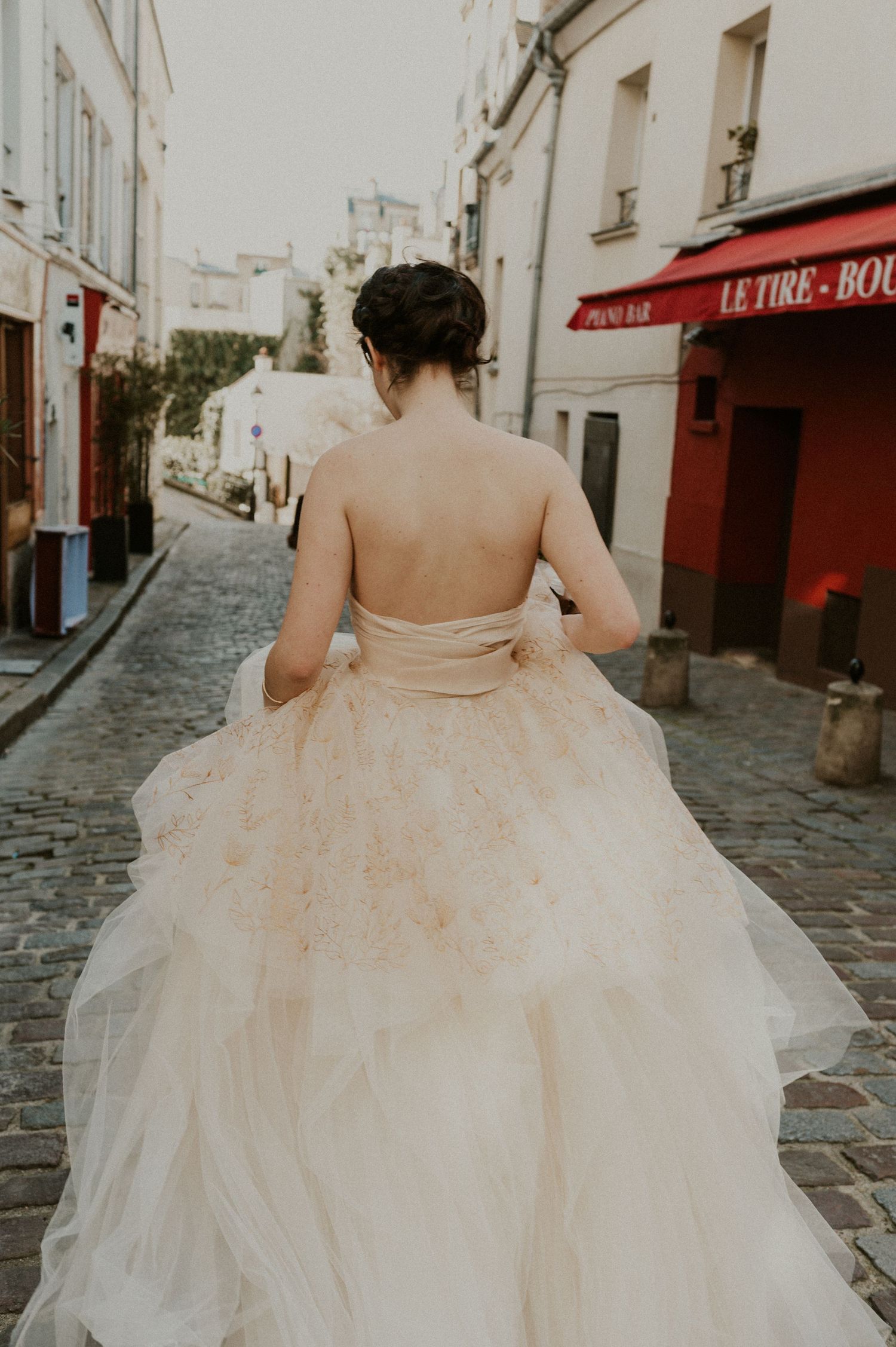 Bride in strapless champagne tulle gown walks down cobblestone street in Paris with red storefront visible.