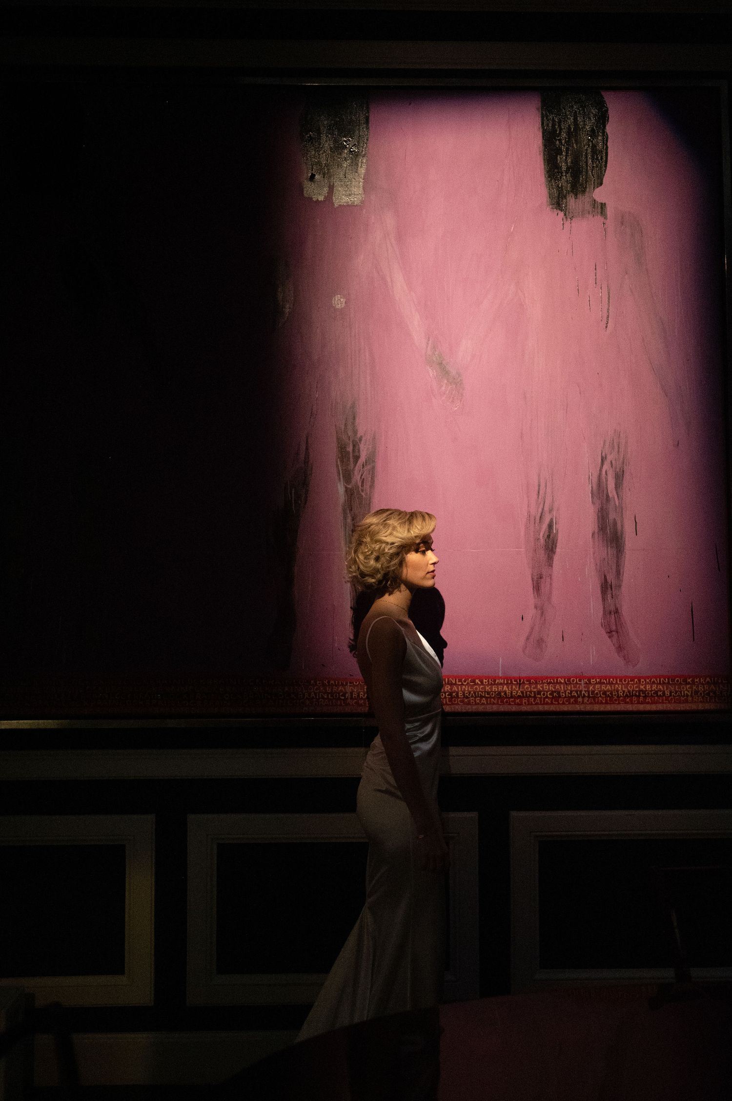 Figure in elegant dress stands in profile against pink textured wall in dark room.