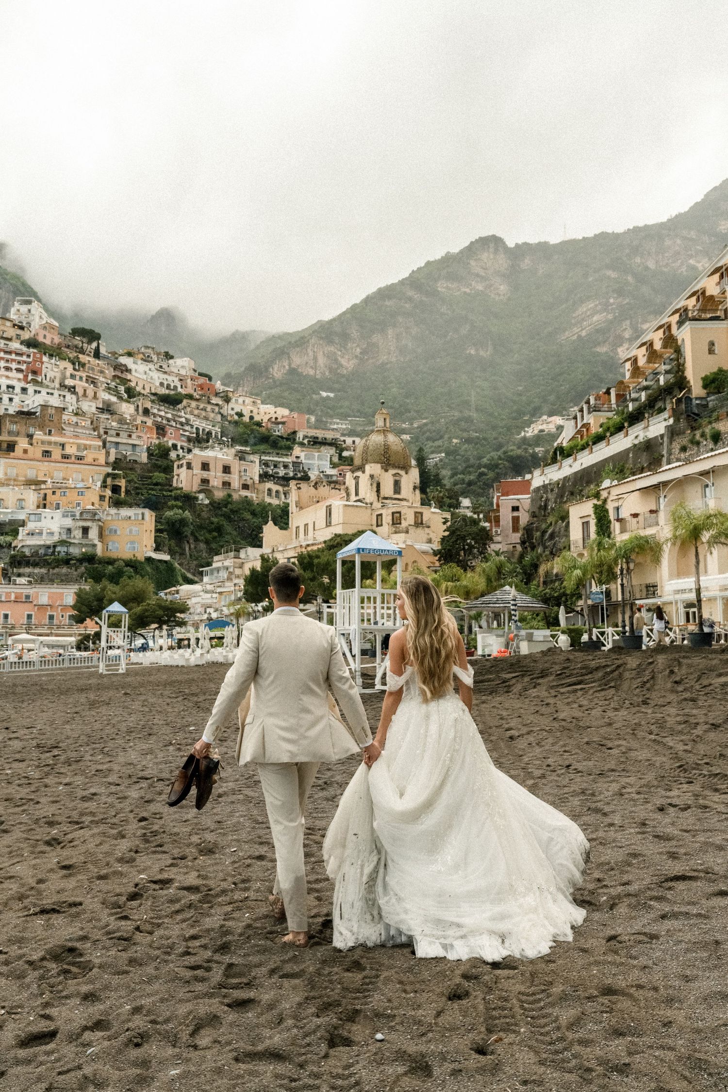 A couple in wedding attire walks hand in hand on a beach with picturesque coastal town views.