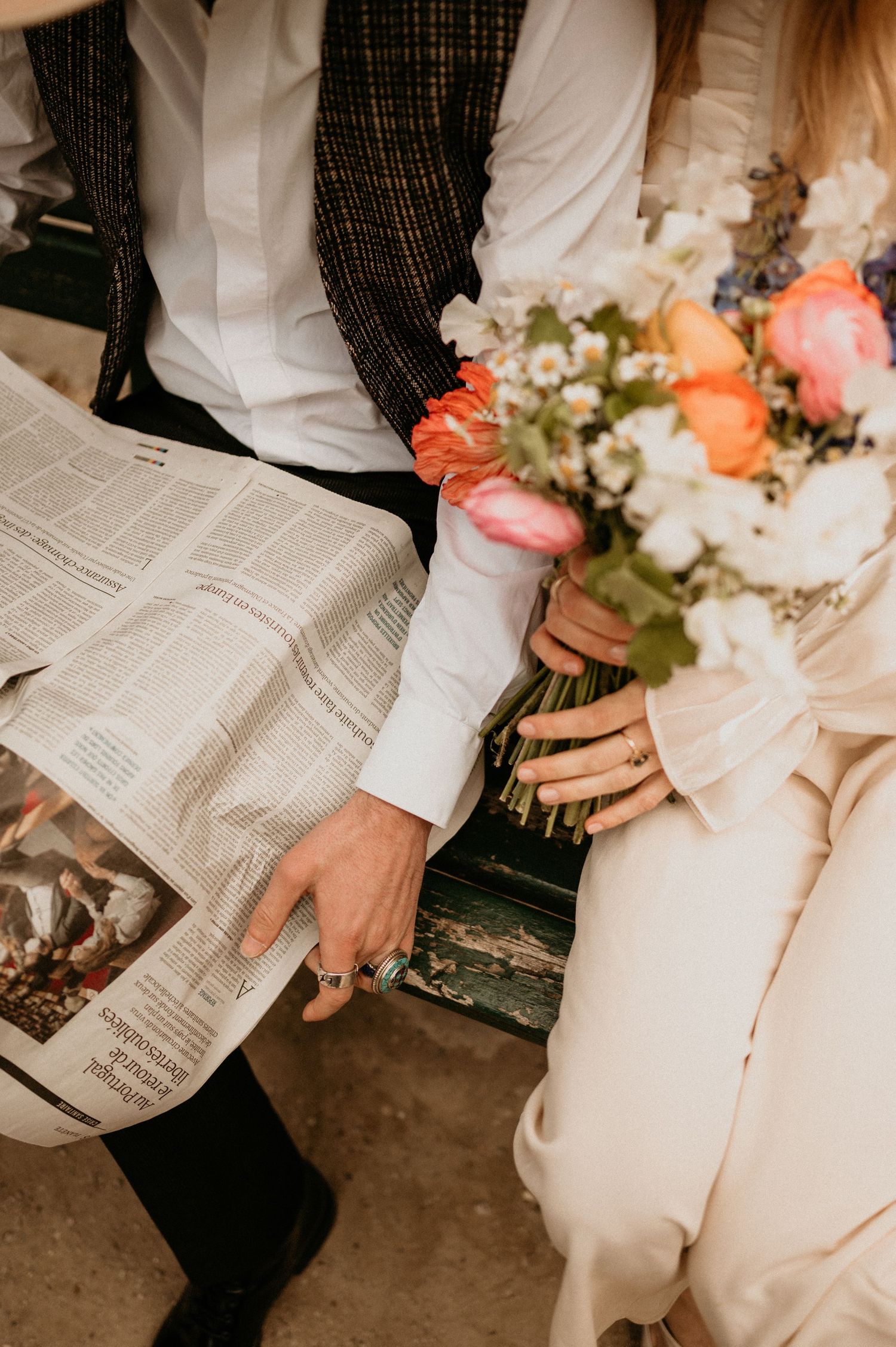 Detailed view of a couple sharing a newspaper and holding a colorful bouquet on a wooden bench.