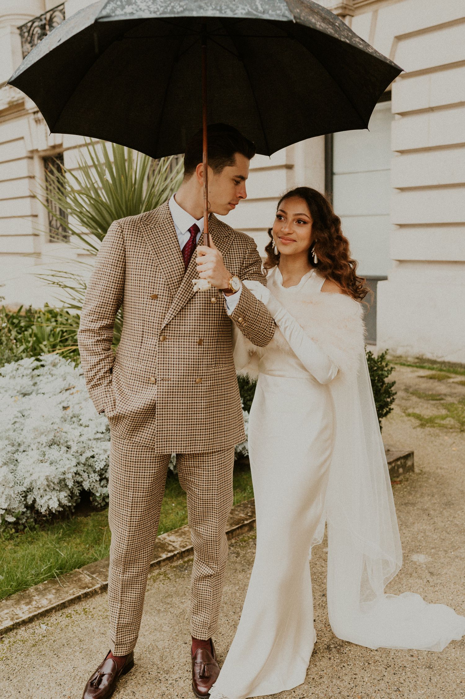 Elegant couple in vintage attire sharing a black umbrella against a white building with blooming gardens.