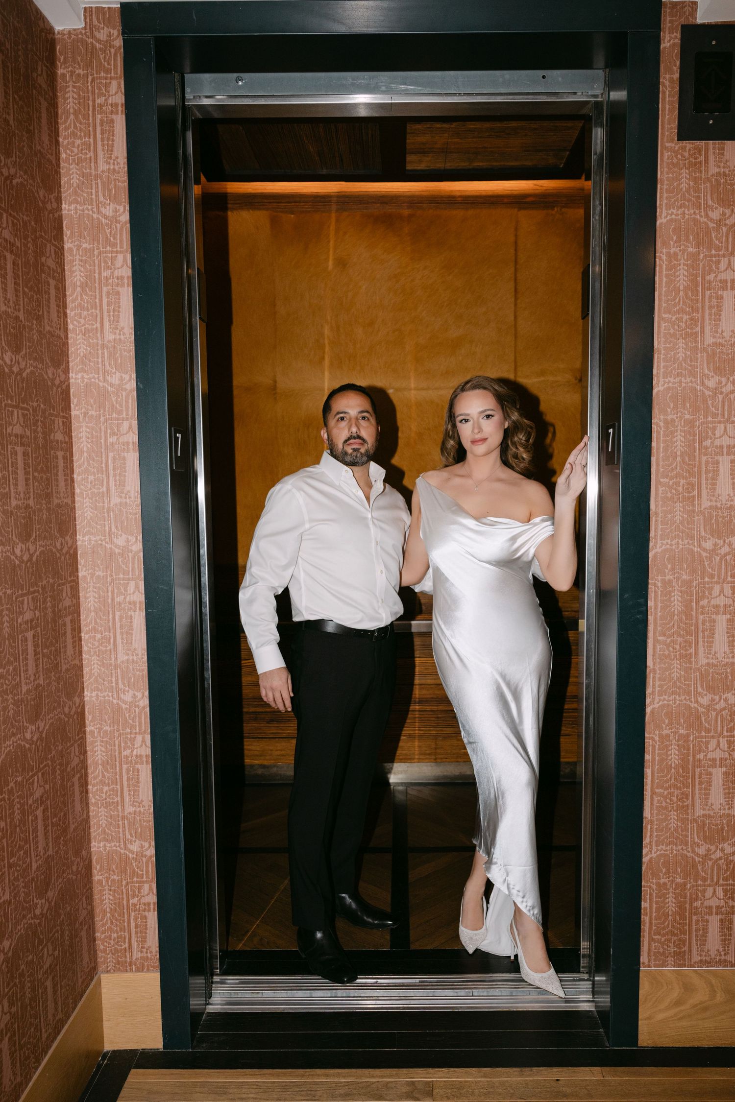 Bride in white satin gown and groom in white shirt stepping out of golden elevator together.