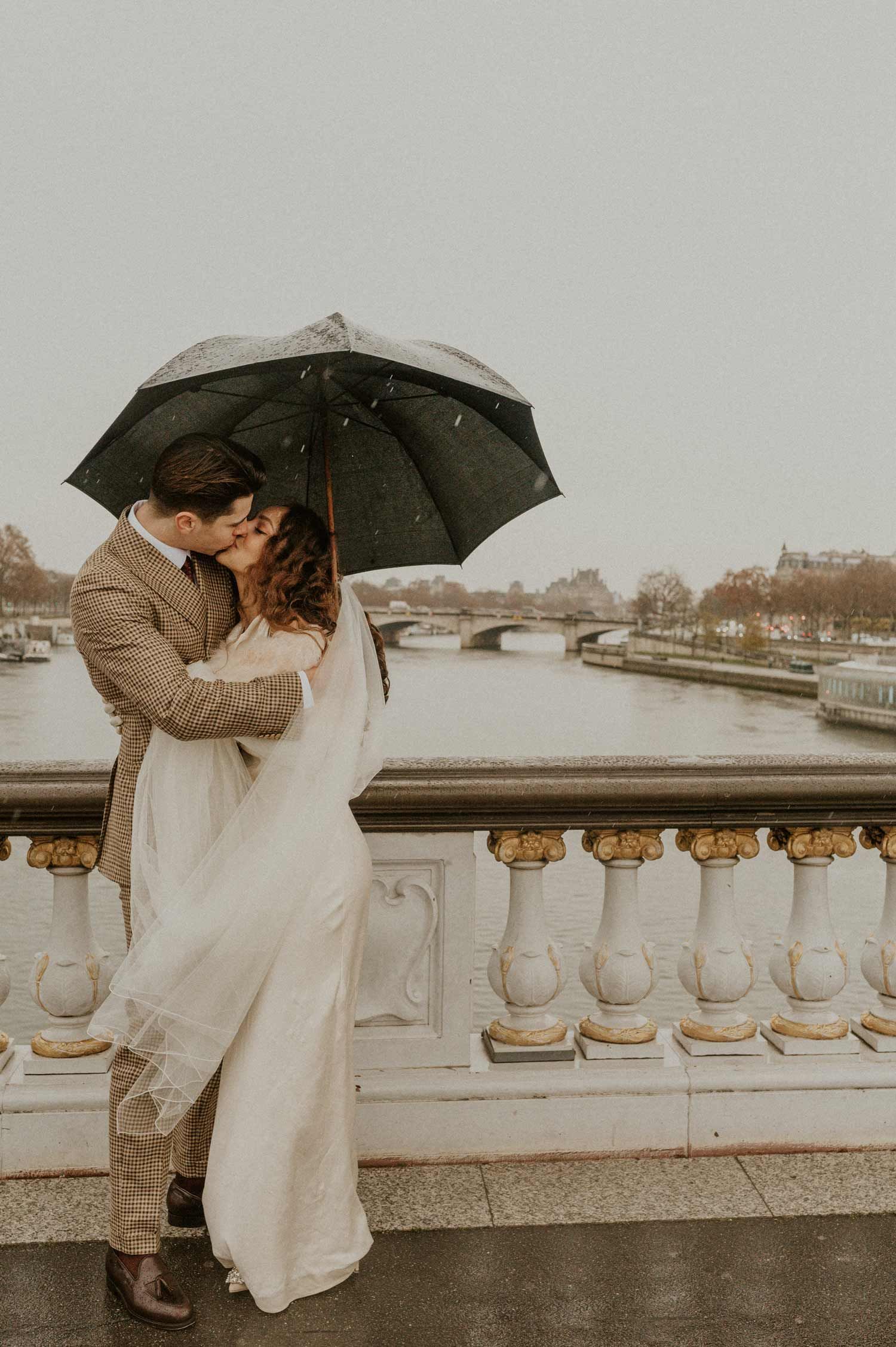 An intimate wedding moment shared under a black umbrella on a historic stone bridge.