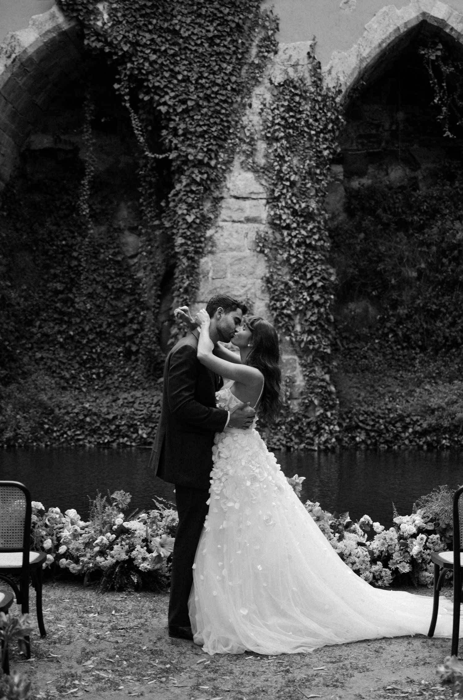 Black and white photo of bride and groom sharing first kiss at ivy-covered stone arch wedding venue.