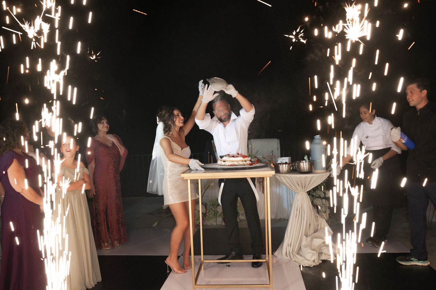 Bride and groom cutting wedding cake surrounded by dramatic sparkler fountains at night.