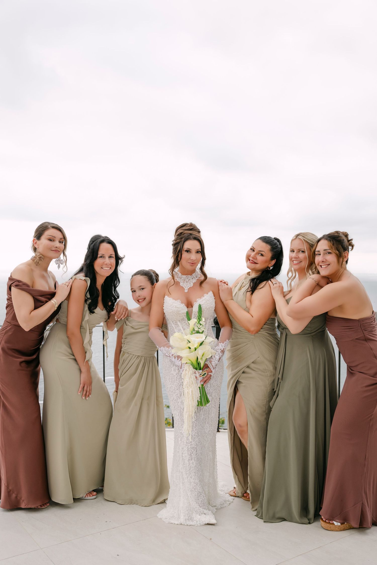 Bride in white lace gown holds bouquet surrounded by six bridesmaids in sage green and brown dresses outdoors.